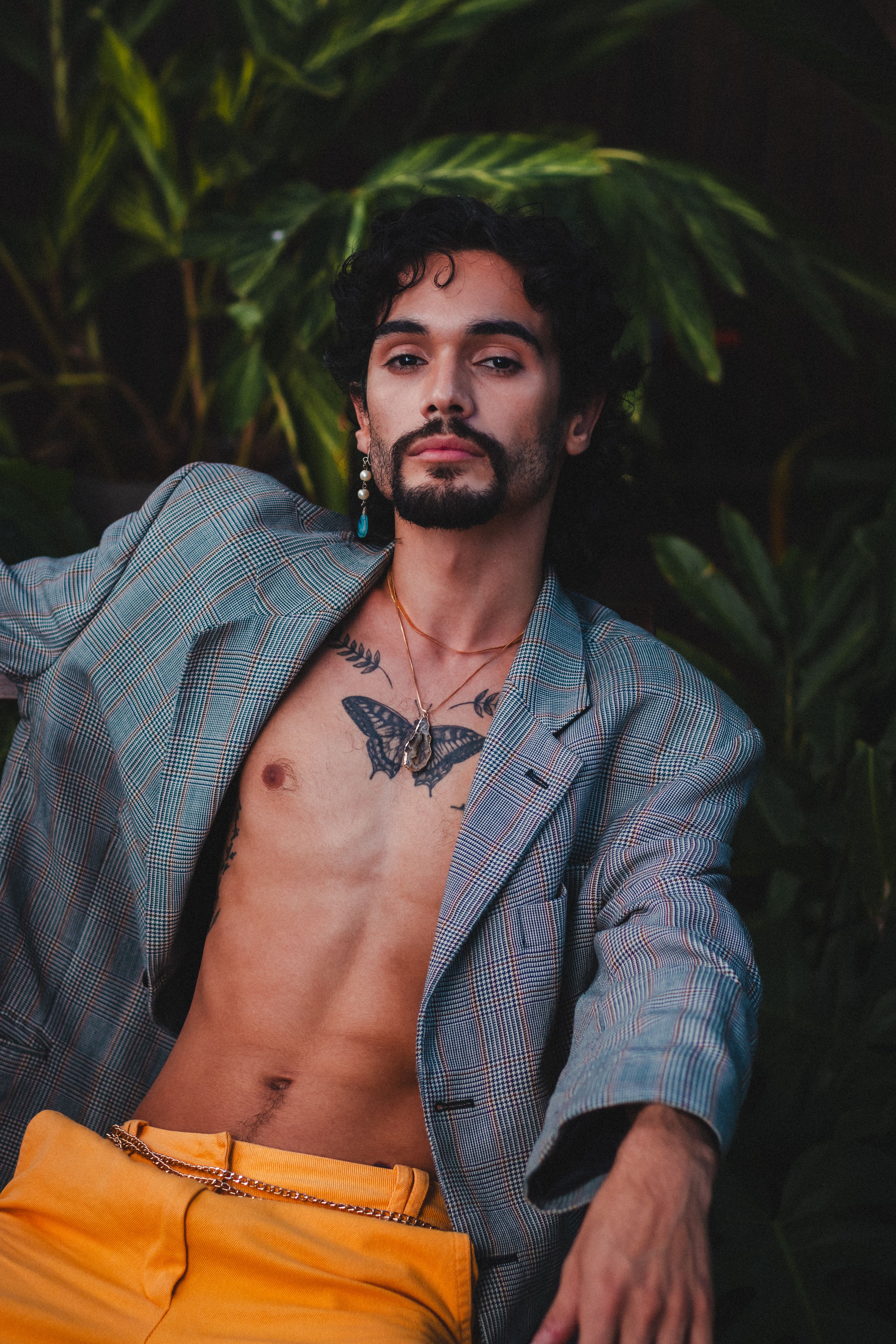 A fashionable portrait of a man with a tattoo, wearing a blazer in a lush outdoor setting.