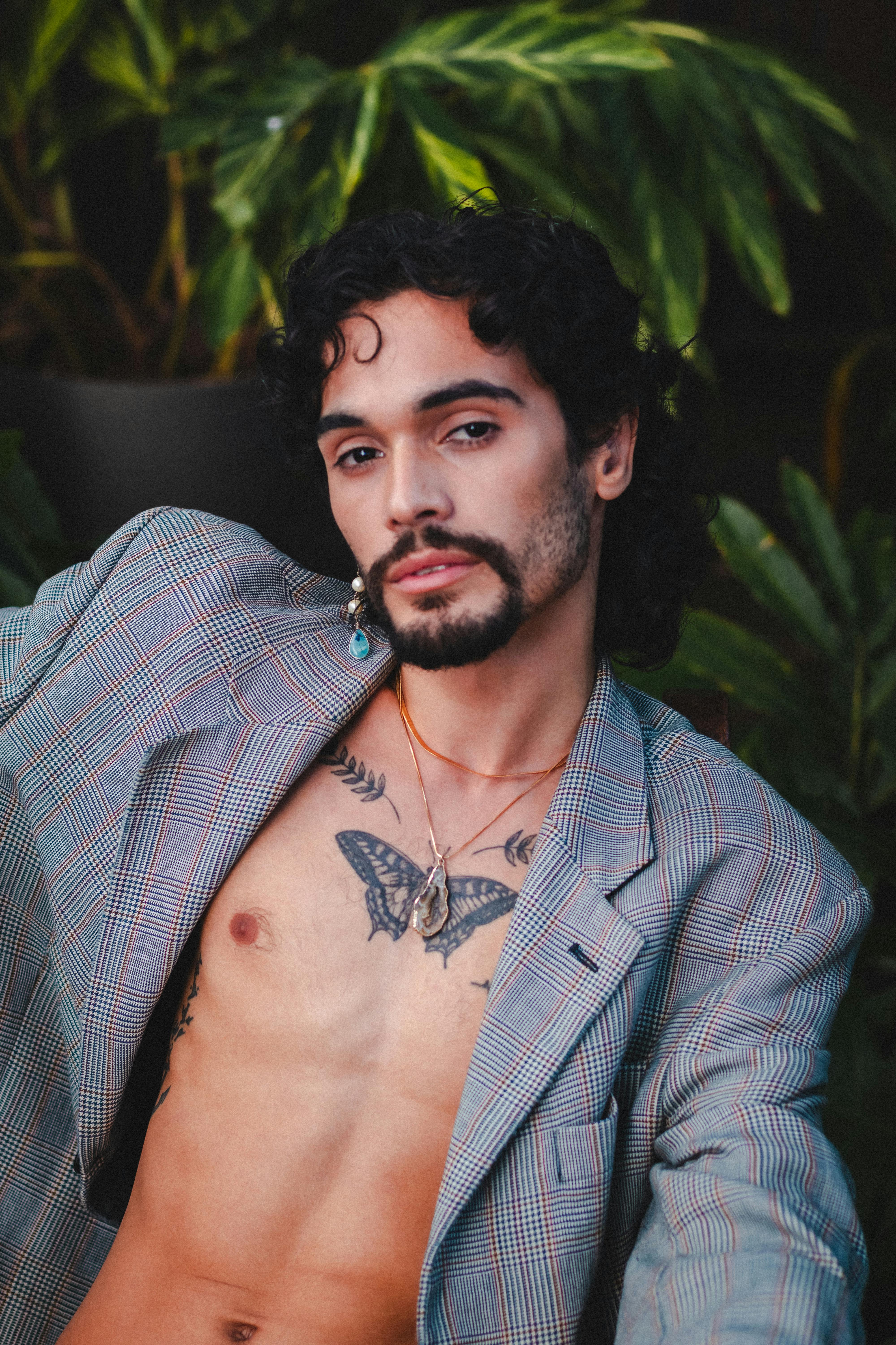 A fashionable, tattooed man in a blazer poses confidently against a lush green background.