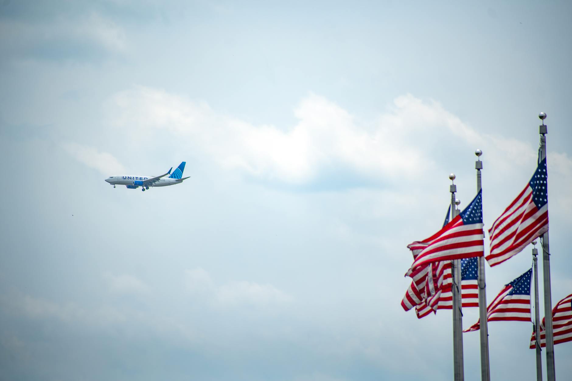 Airplane flying near waving American flags against a bright blue sky, symbolizing travel and patriotism.
