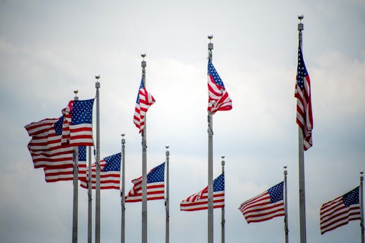 A row of American flags waving proudly at the base of the Washington Monument against a cloudy sky.