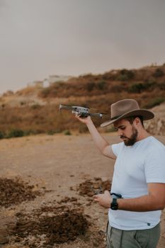 Man with hat controlling a drone over a desert landscape during the day.