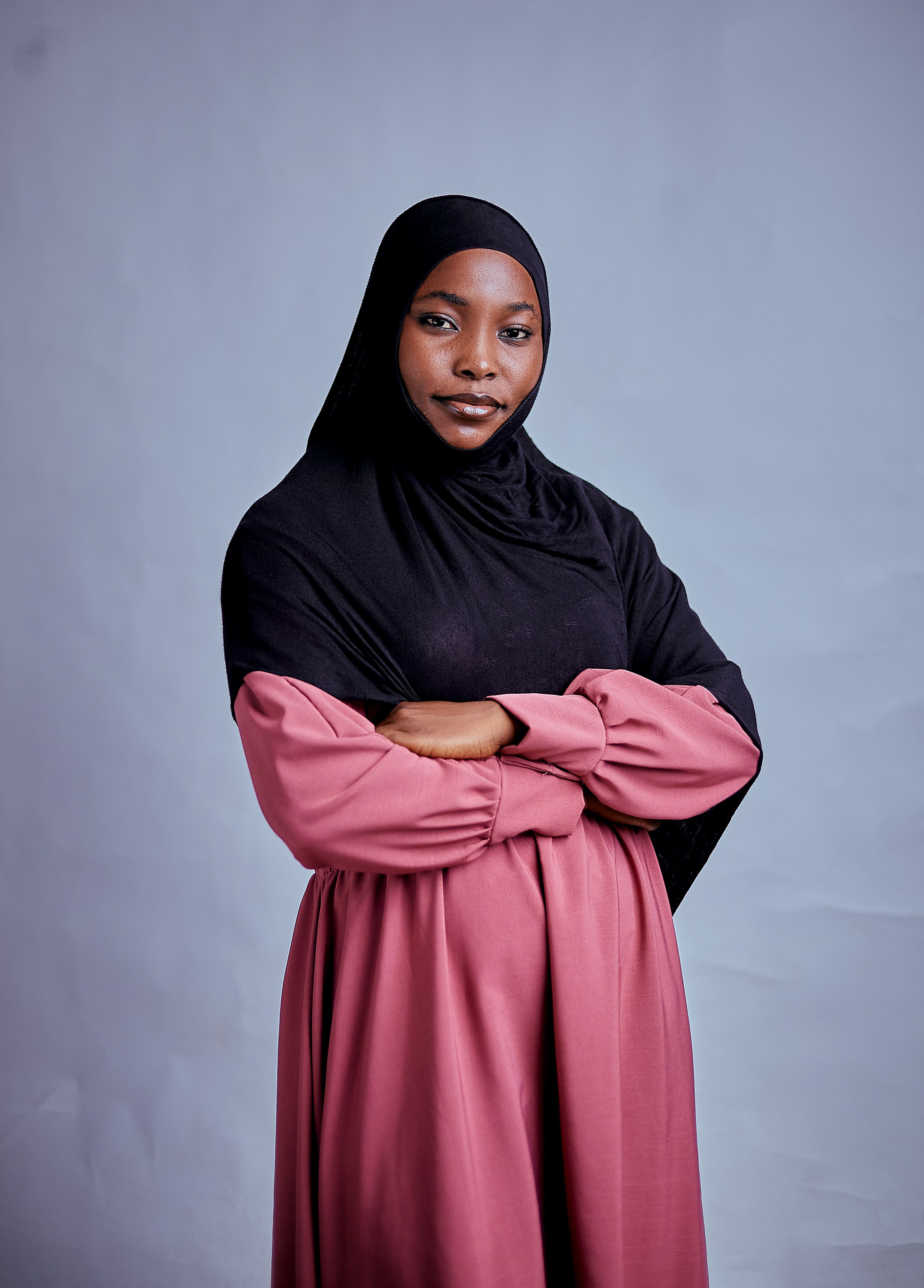 A confident woman wearing a hijab and pink dress poses with arms crossed.