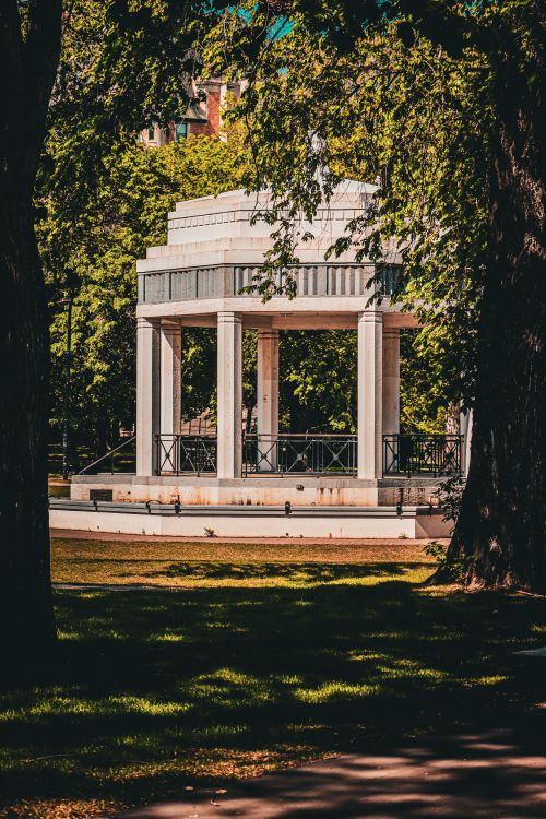 Free A white gazebo nestled in a serene park setting surrounded by verdant trees during a bright summer day. Stock Photo