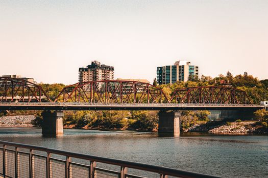 Steel bridge spanning river with urban skyline and trees, under a clear daytime sky.