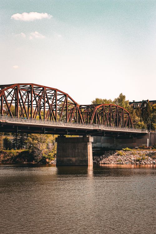 Free A vintage iron bridge spans a tranquil river surrounded by lush greenery in summer. Stock Photo