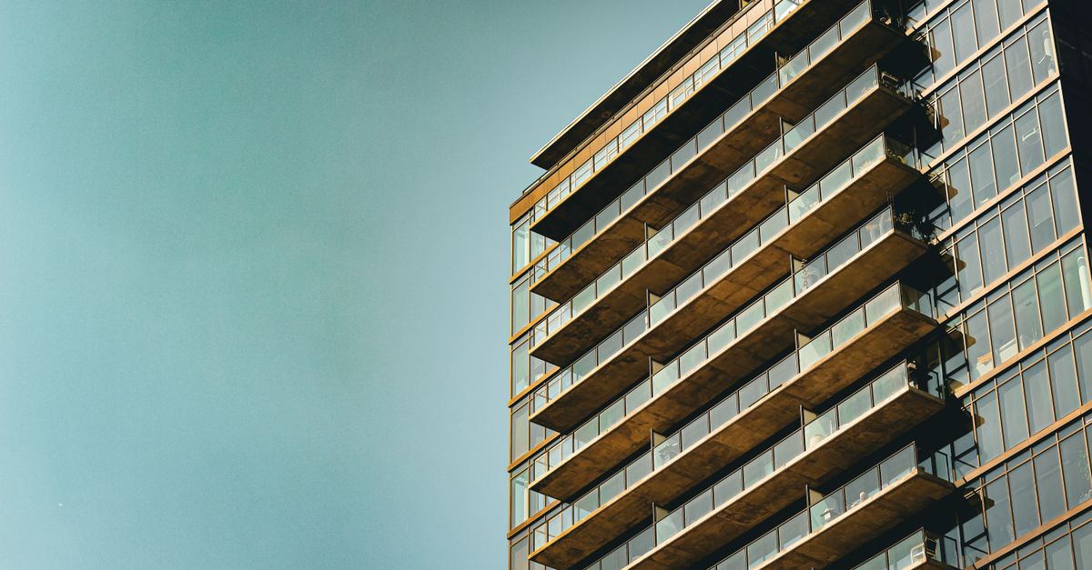 Sleek modern apartment building with glass facade and balconies, set against a clear blue sky.