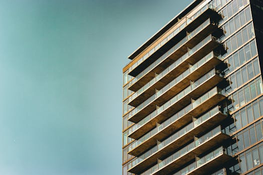 Sleek modern apartment building with glass facade and balconies, set against a clear blue sky.