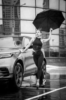 Stylish woman poses with umbrella by a luxury car in a rainy urban setting, black and white.