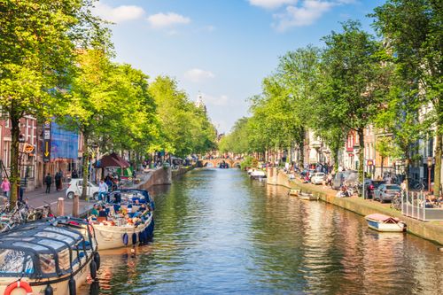 Free Vibrant Amsterdam canal view with boats and lush greenery under a clear blue sky, perfect for travel insights. Stock Photo