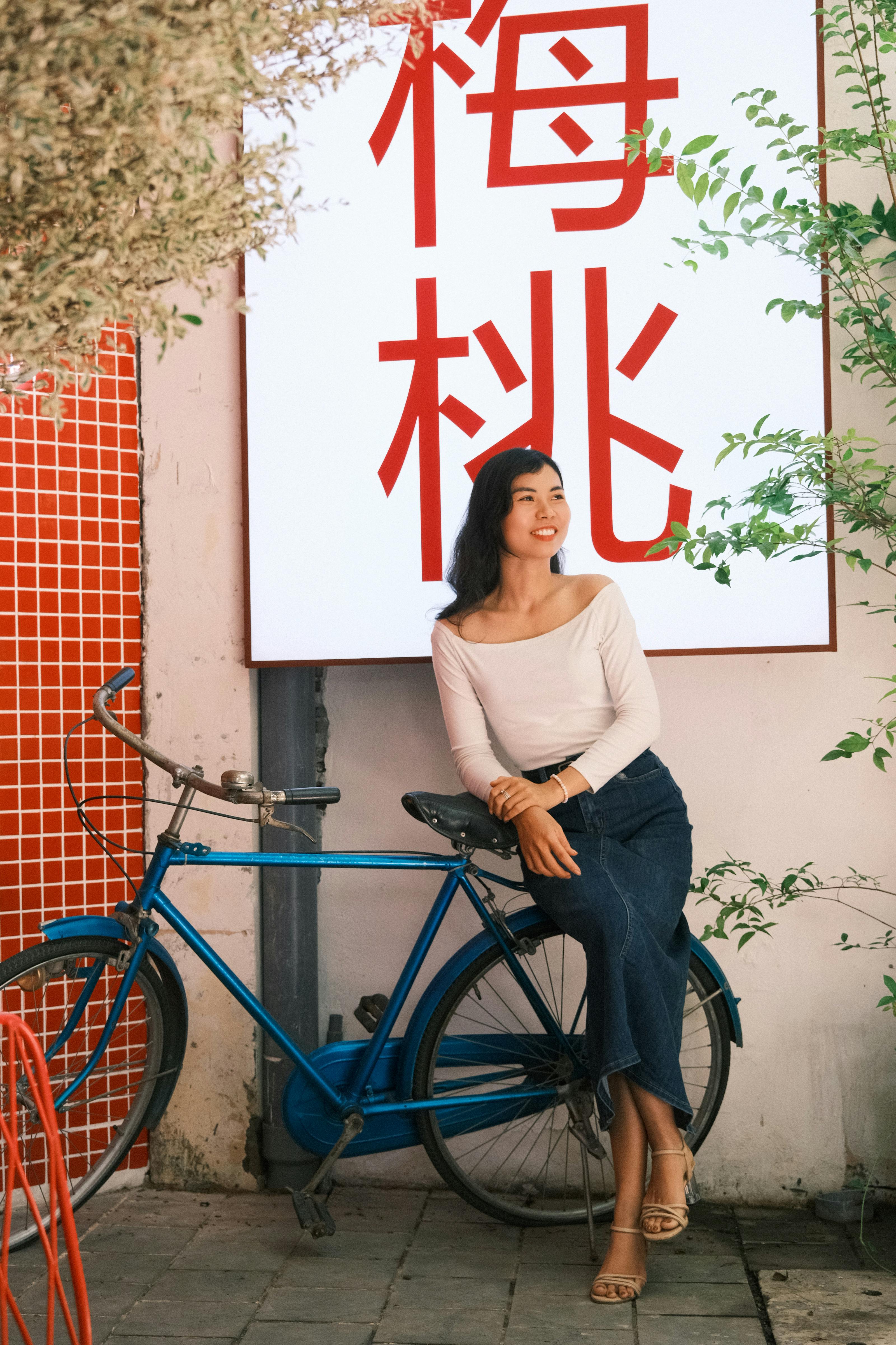 Smiling woman sits by a blue bicycle near plants in urban area.