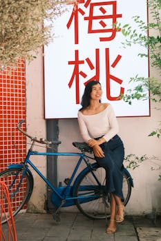 Smiling woman sits by a blue bicycle near plants in urban area.