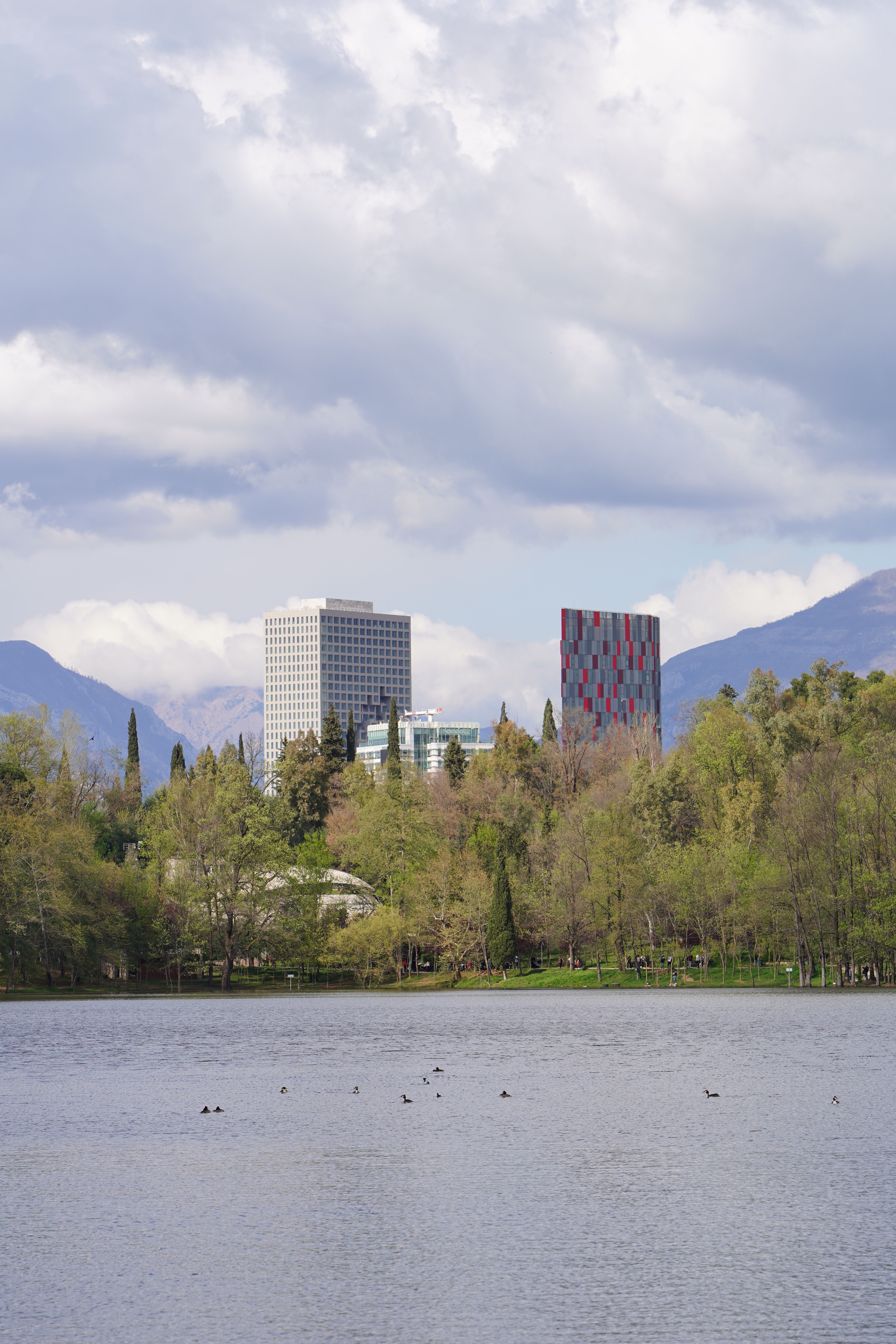 Serene lake with urban skyline, lush trees, and distant mountains beneath a cloudy sky.