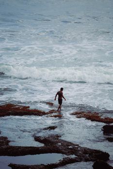 A solitary man walks along the rocky coastline of Bali, Indonesia, surrounded by blue ocean waves.