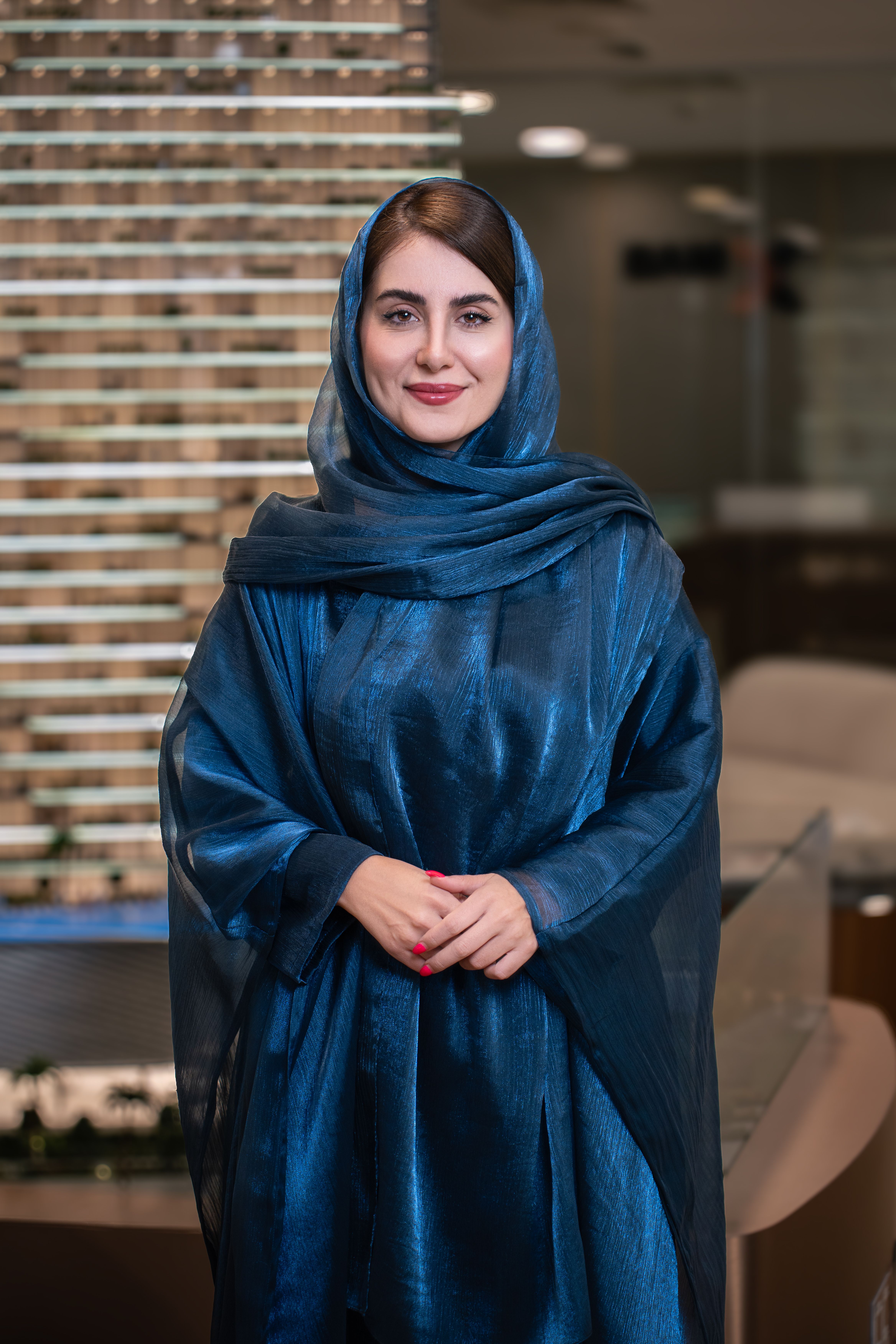 Elegant portrait of a woman in a blue hijab standing indoors with a confident smile.