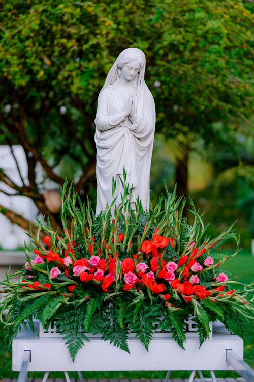 Free Statue of Virgin Mary adorned with vibrant red and pink flowers in a serene garden setting. Stock Photo