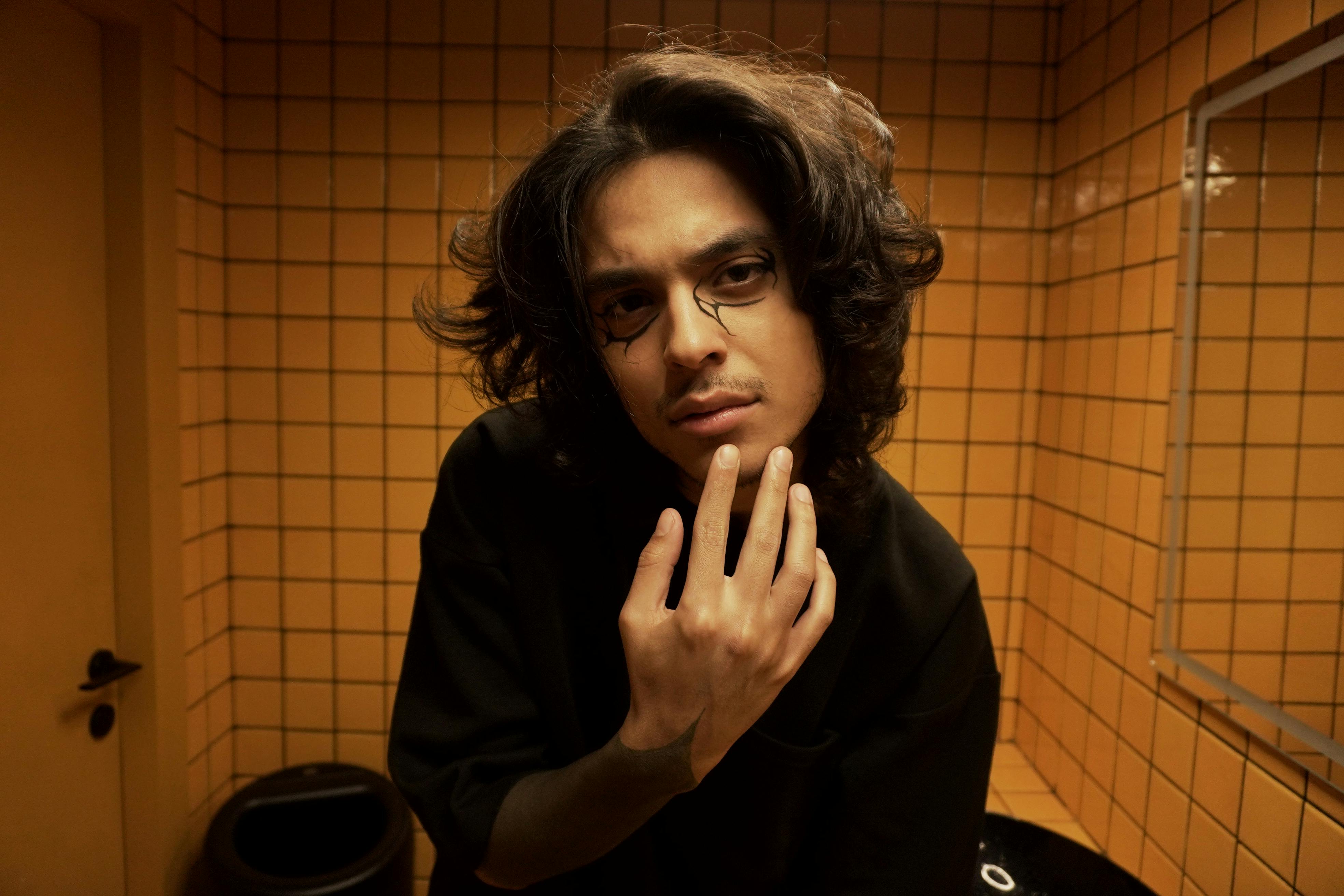 Young man with artistic makeup poses thoughtfully in a tiled room.