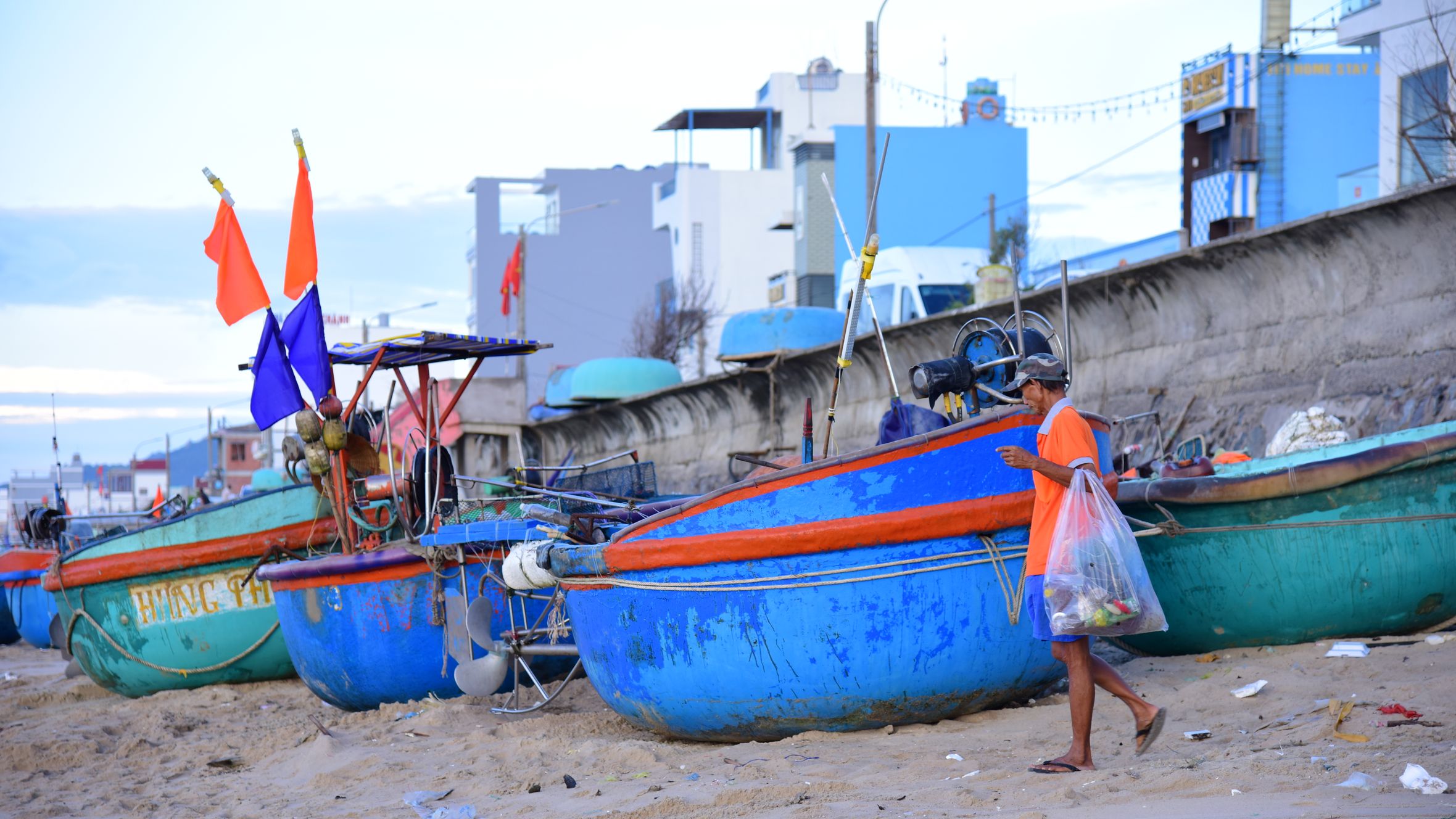 A fisherman carries goods past colorful fishing boats on a sandy beach, showcasing coastal livelihood.