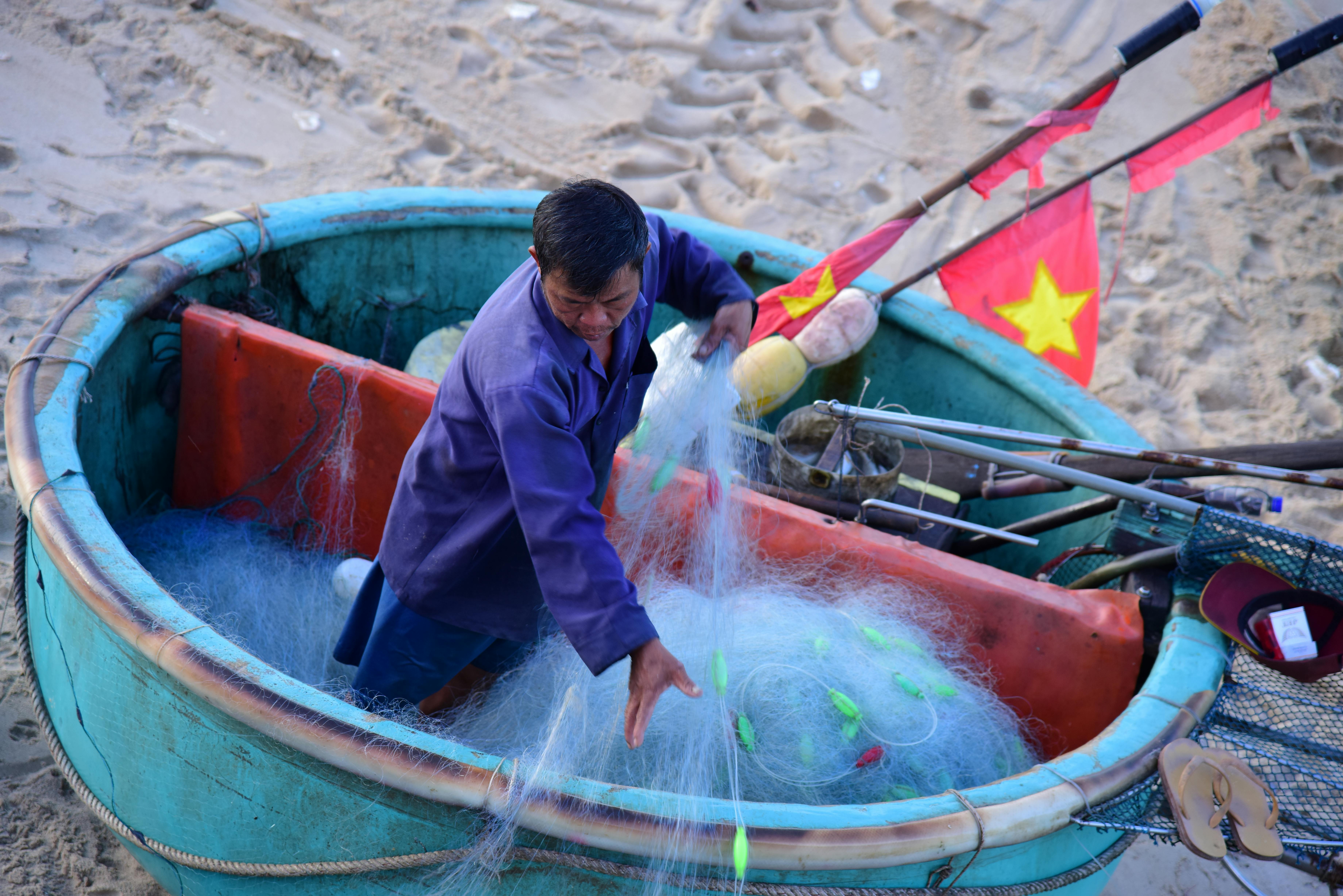 A fisherman arranges nets in a traditional round Vietnamese basket boat on a sandy beach.