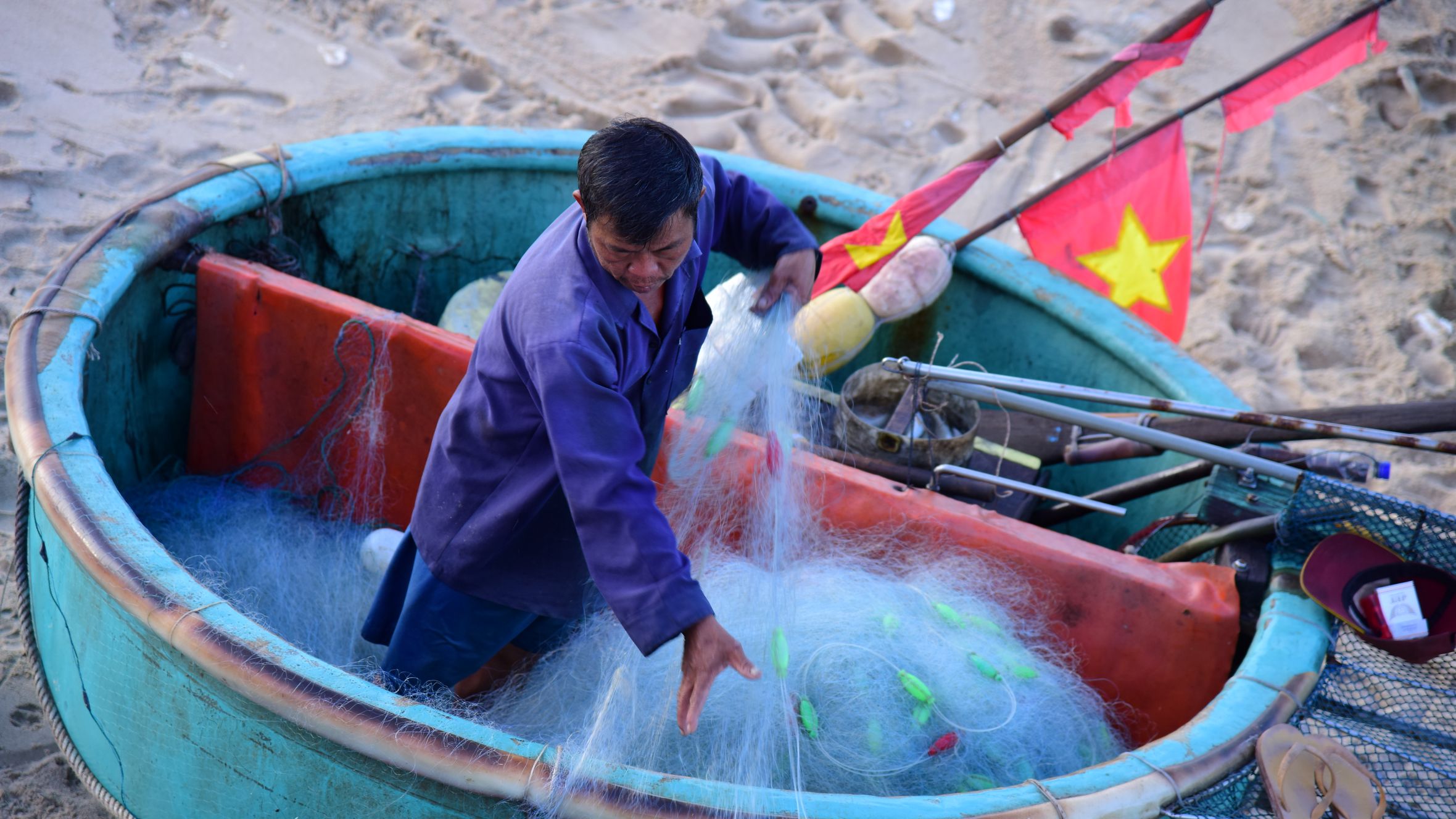 A fisherman arranges nets in a traditional round Vietnamese basket boat on a sandy beach.
