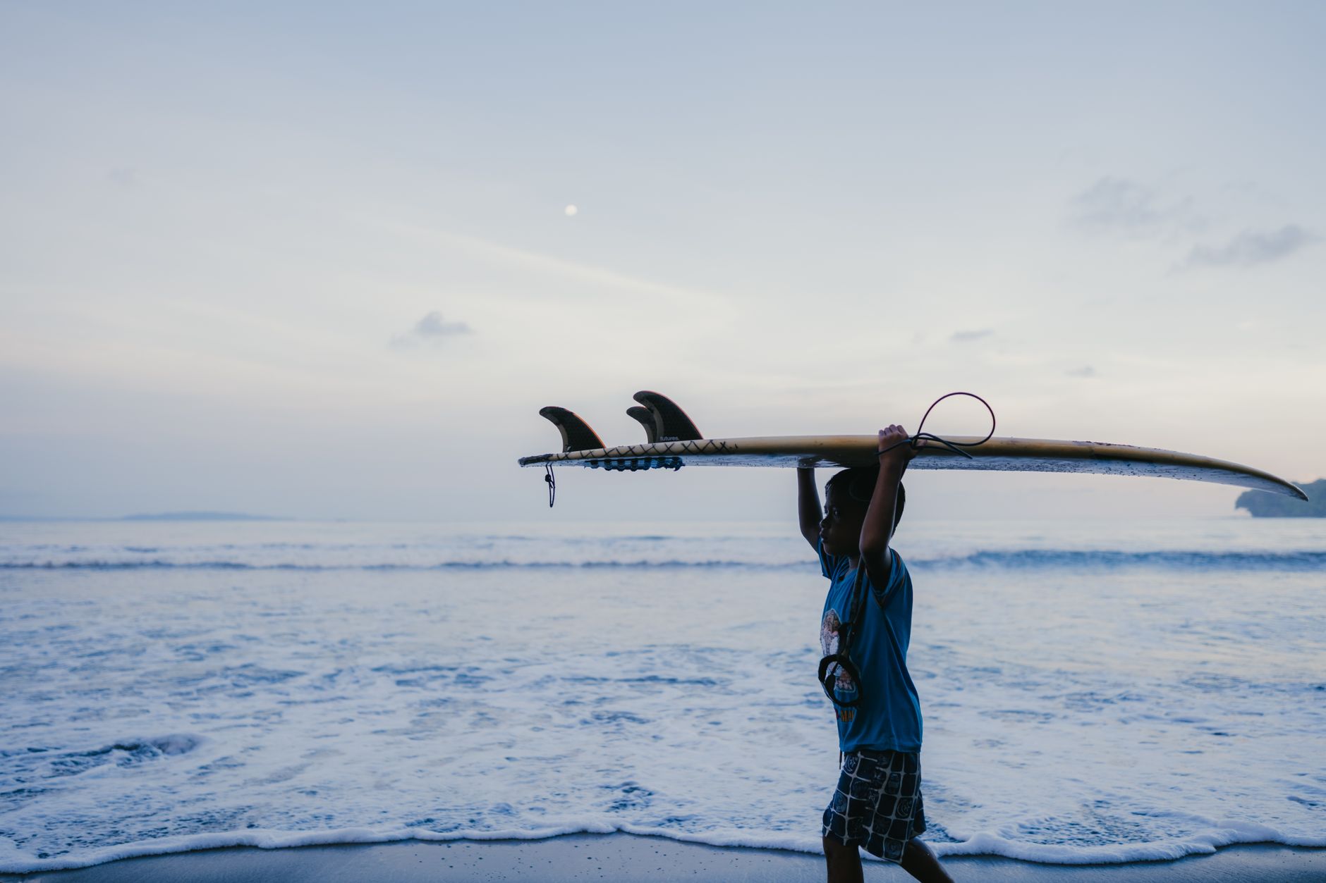 A surfer carrying a board along the beach at dusk in Jawa Barat, Indonesia.
