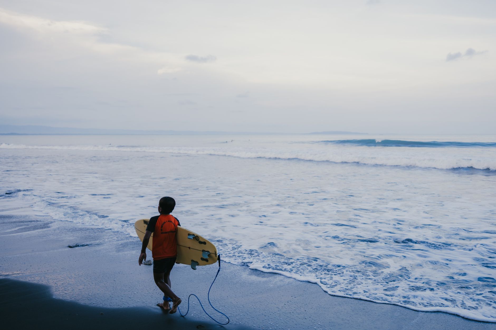 A young surfer with a surfboard walks along a serene Indonesian beach at dawn, ready for adventure.