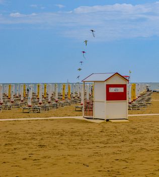 A peaceful sunny beach with a lifeguard hut, empty loungers, and colorful kites in the sky.