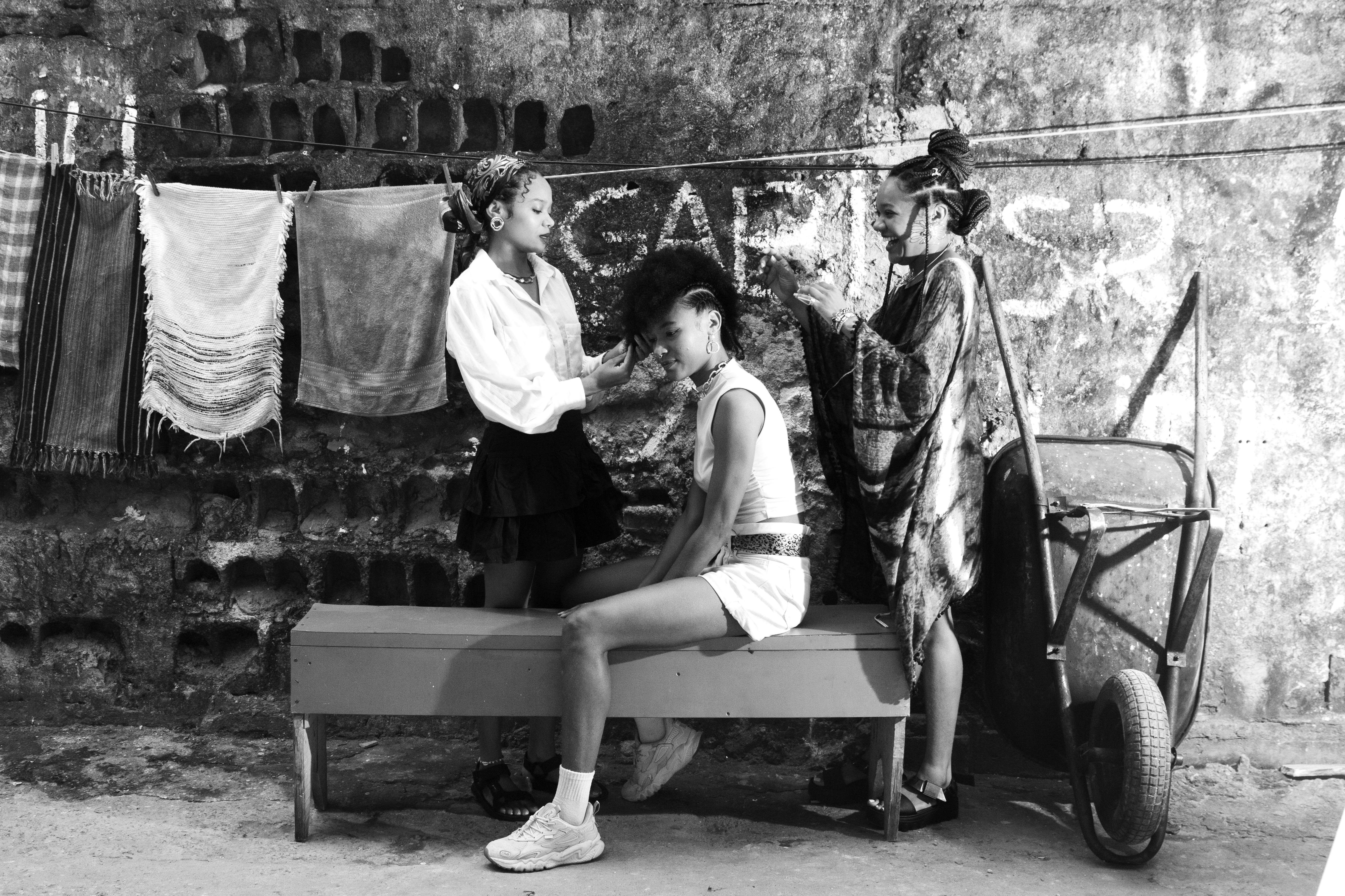 Black and white photo of street hair braiding with three women, urban wall background.