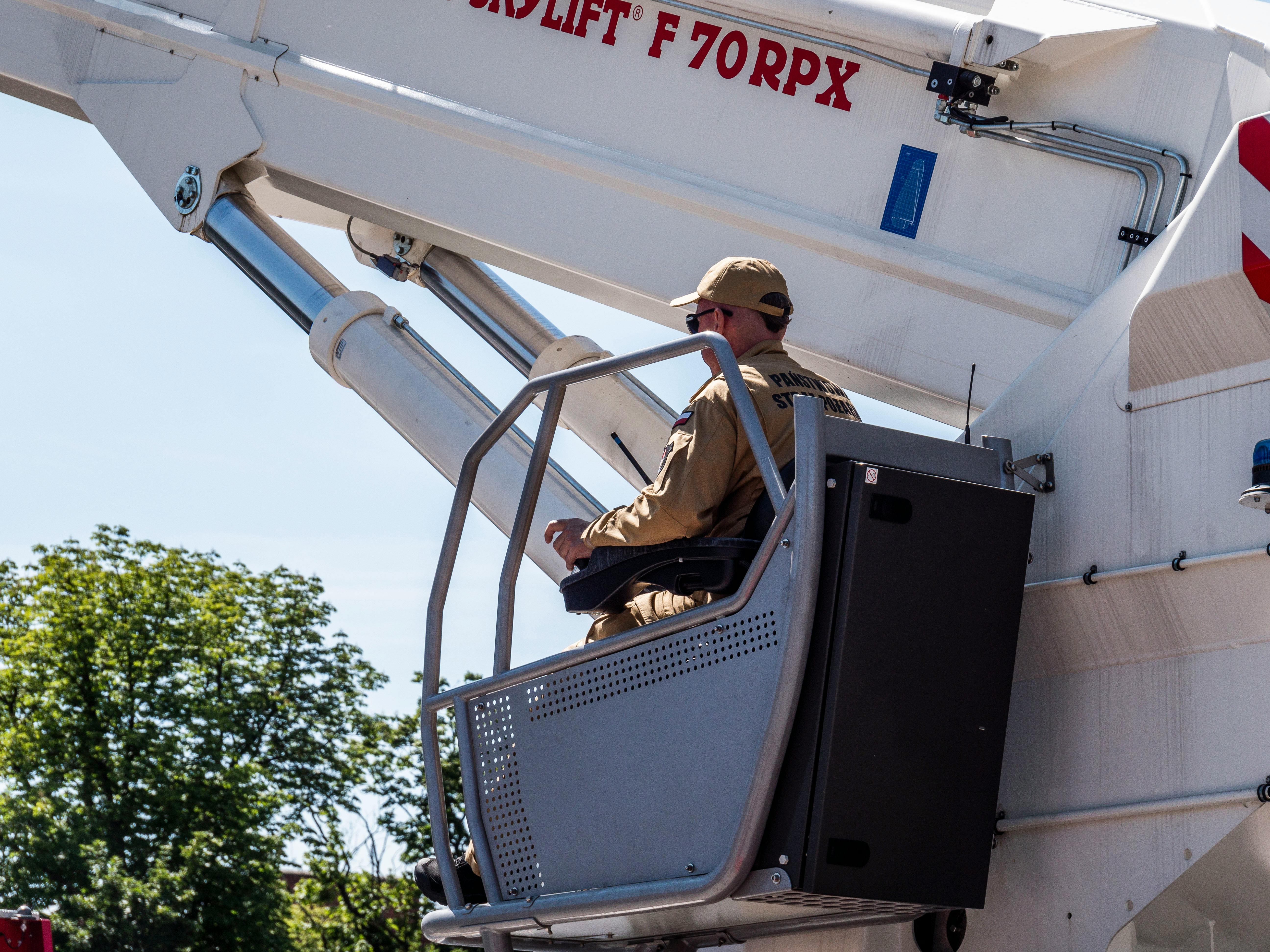 A person operates a crane basket with a control panel. They wear a tan uniform and sunglasses, and the crane is white.