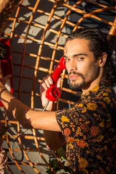 A fashionable young adult man with a vintage red telephone in a sunny woven booth.