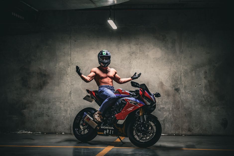 Shirtless motorcyclist posing confidently on a red sports bike in a dimly lit indoor setting.