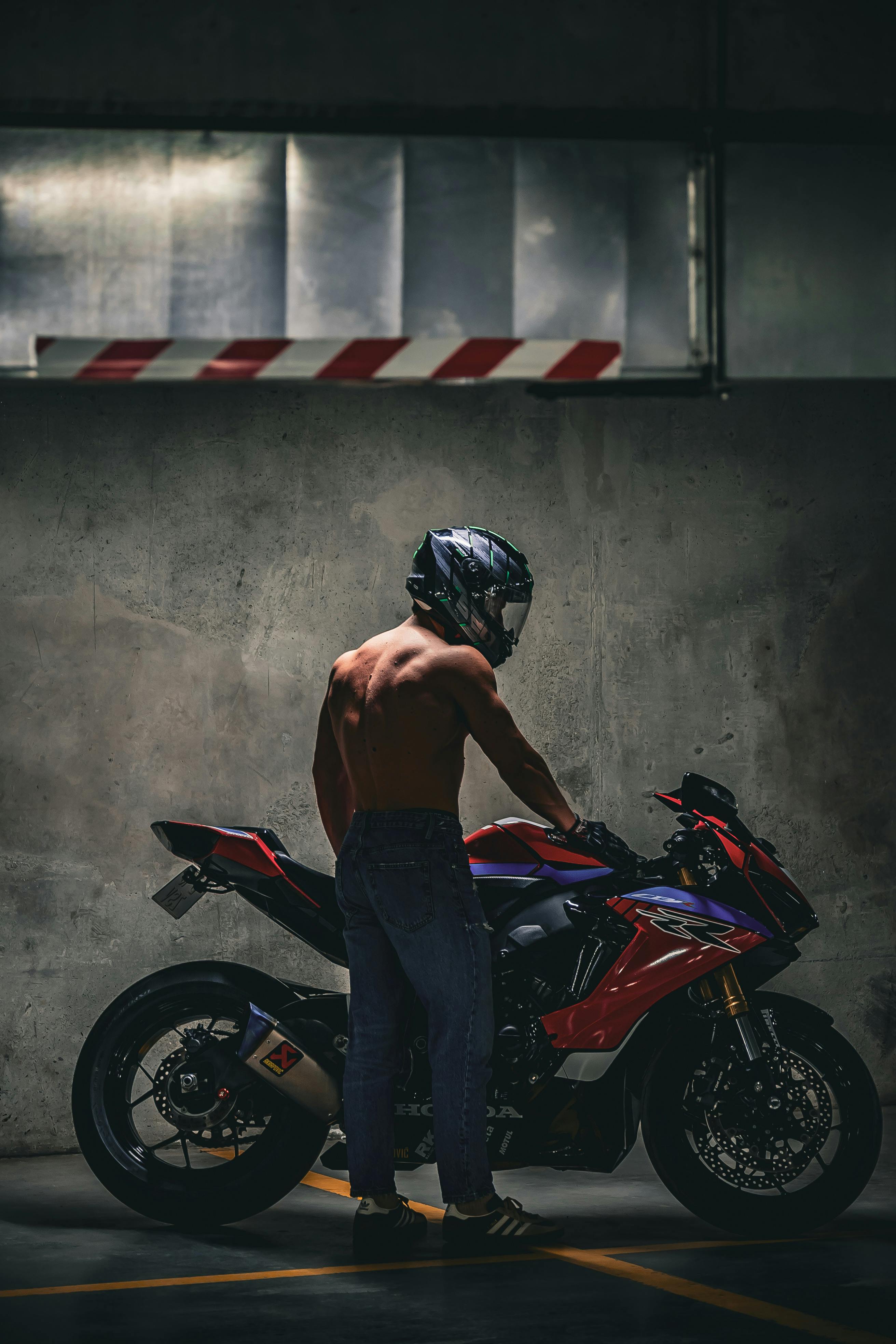 A shirtless man wearing a helmet stands beside a motorcycle in a dimly lit garage.