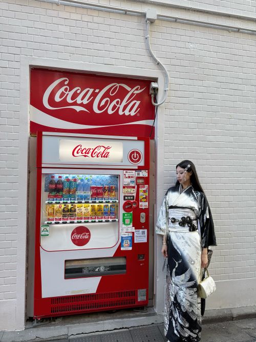Free Asian woman in a kimono stands beside a Coca-Cola vending machine against a white brick wall. Stock Photo