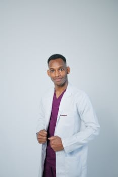 A confident male doctor wearing a white lab coat over maroon scrubs with a gray background.