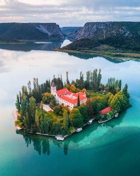 航拍画面中，一座修道院坐落于宁静的岛屿上，四周环绕着晶莹剔透的蓝色海水