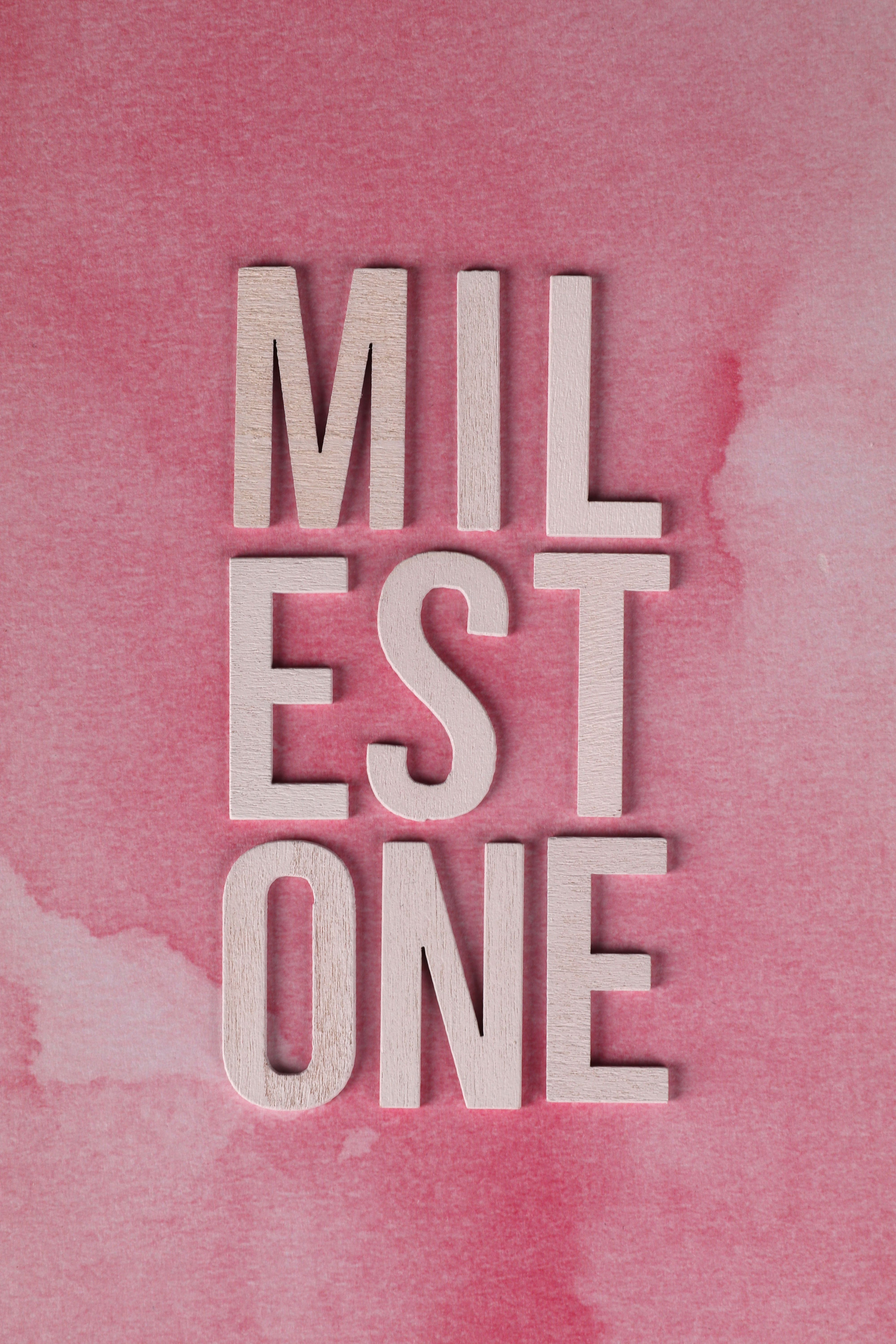Wooden letters spelling 'Milestone' on a pink watercolor backdrop, symbolizing achievement.