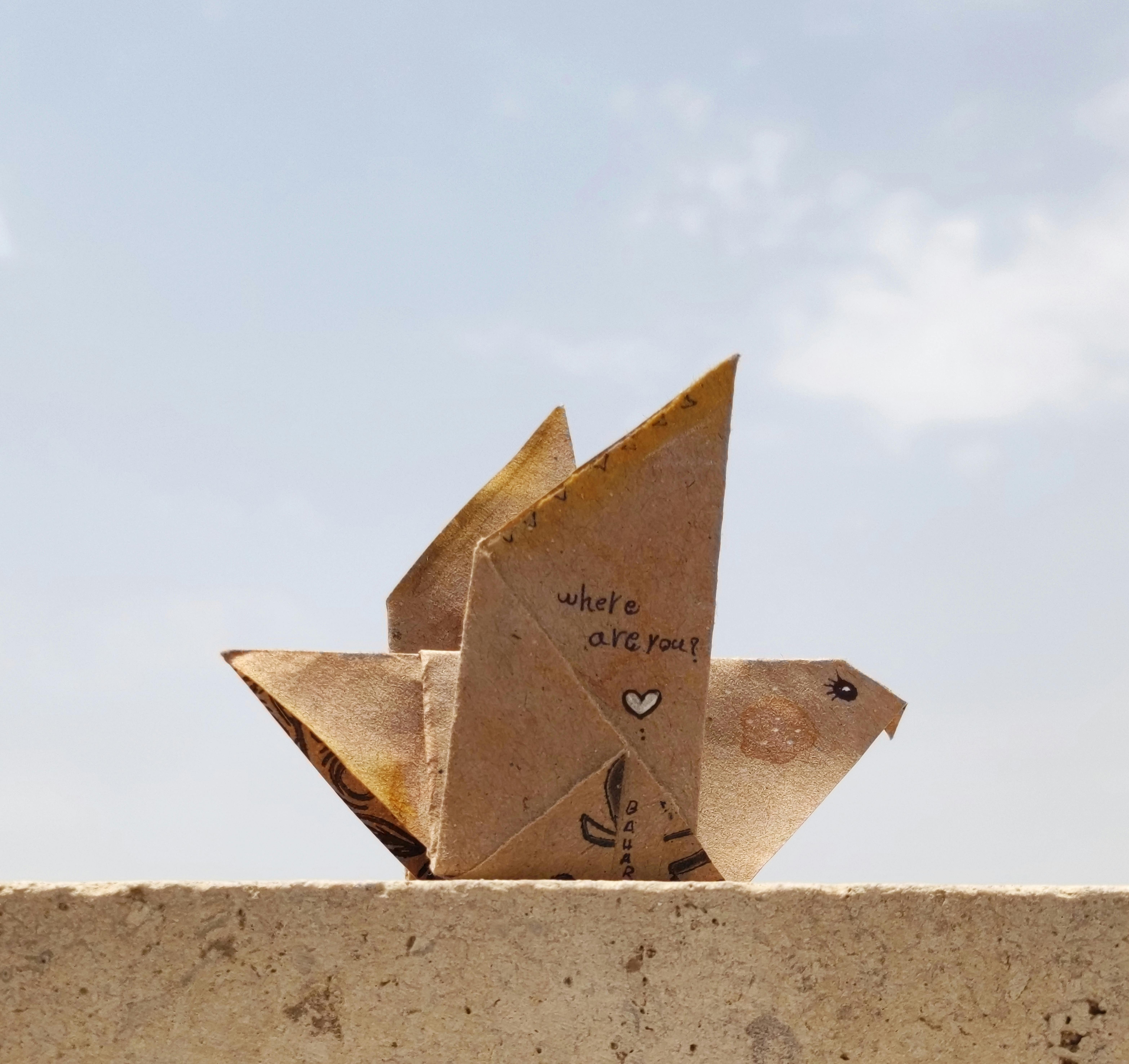 A paper origami bird on a ledge with 'Where are you?' written on it against a clear sky.