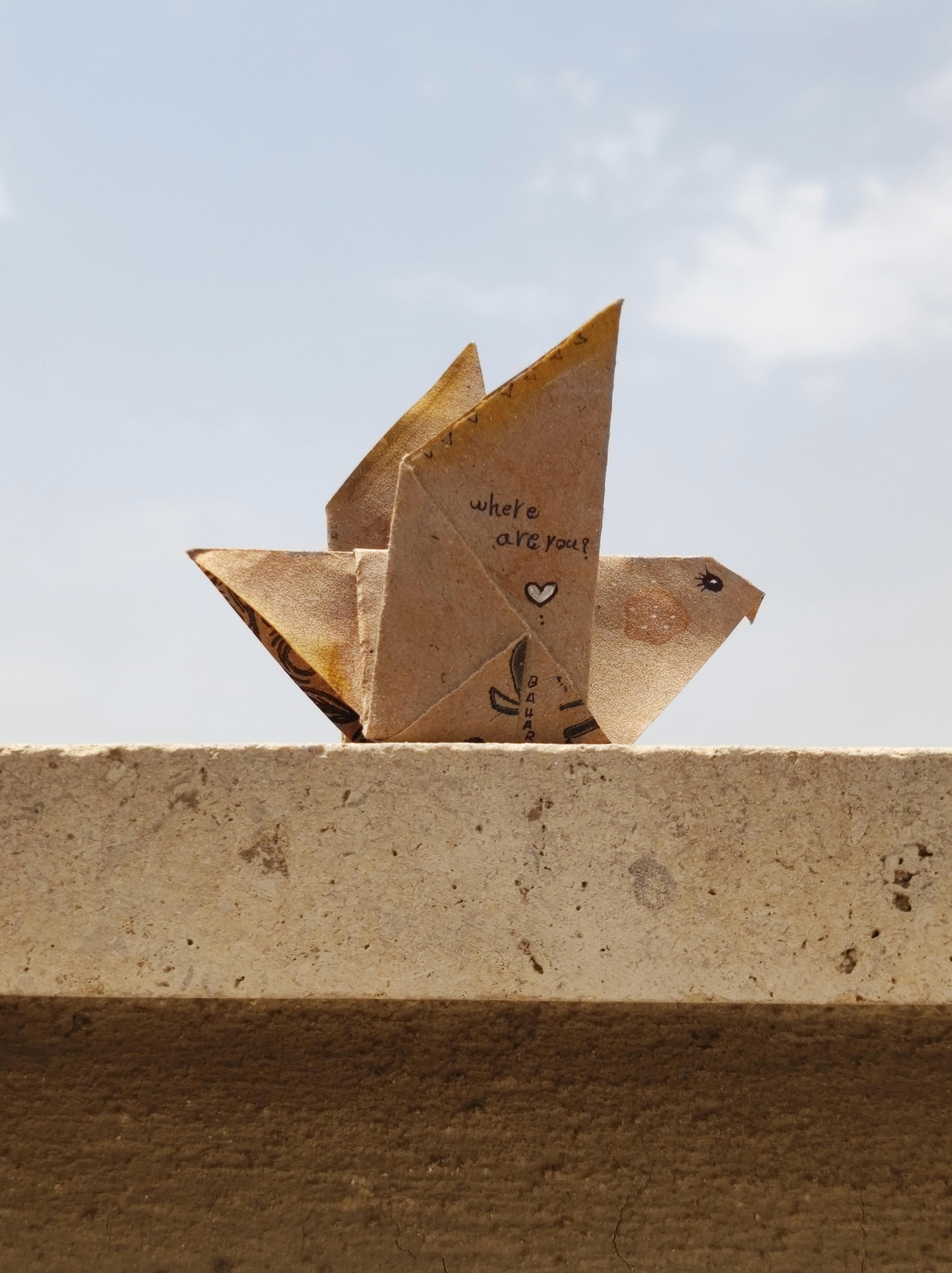 An origami bird with 'where are you?' text sits on a stone ledge under a bright sky.