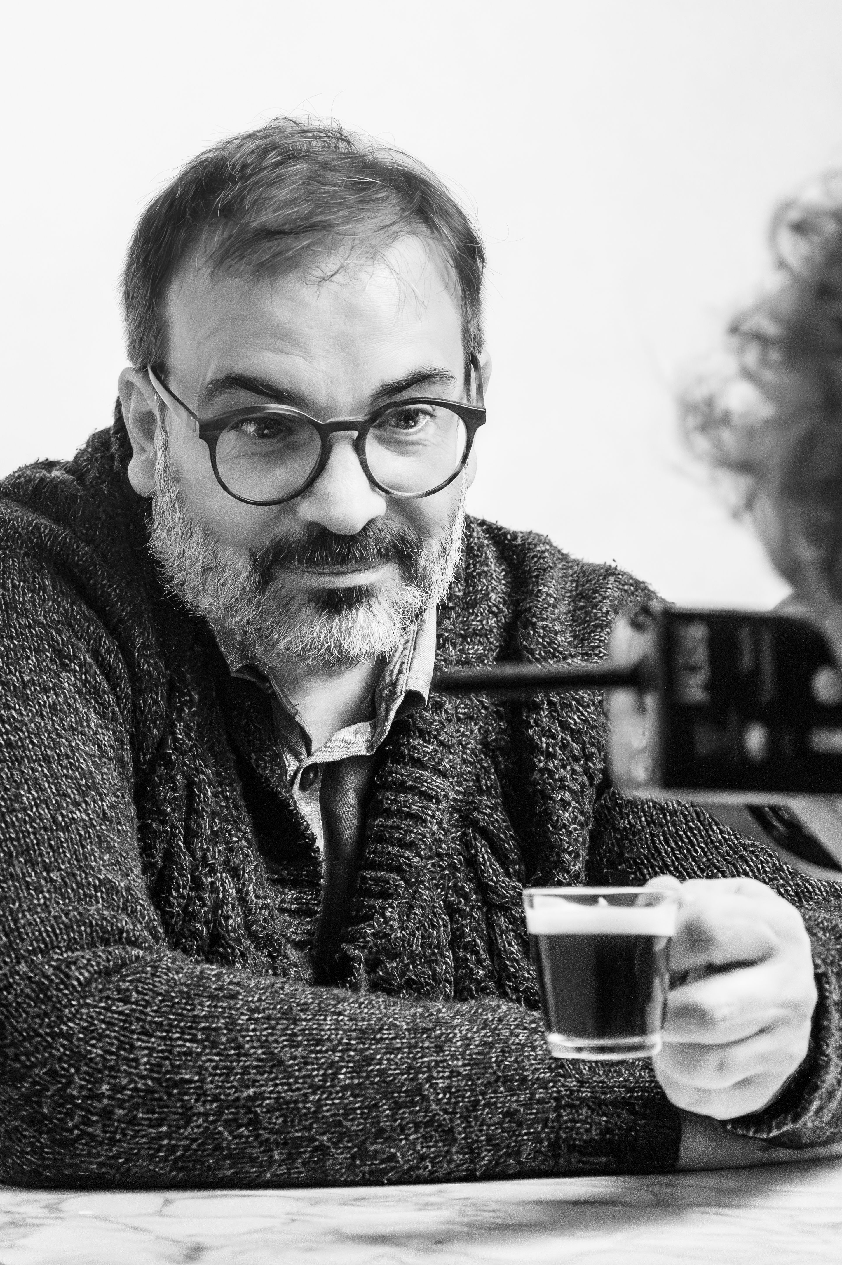 A man with glasses smiles while holding a coffee cup, captured indoors.