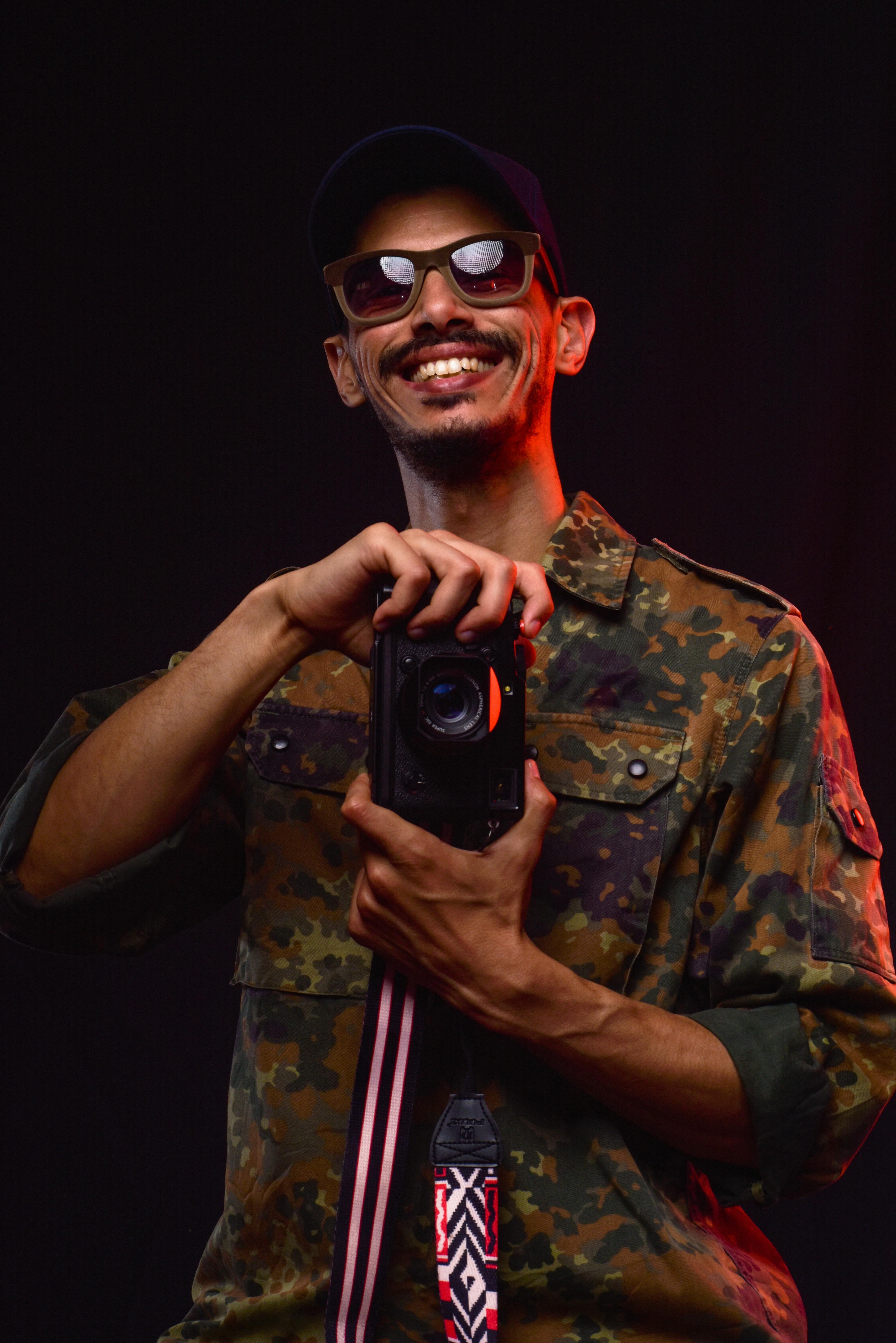 Confident man in camouflage shirt holding a camera, smiling in a studio setting.