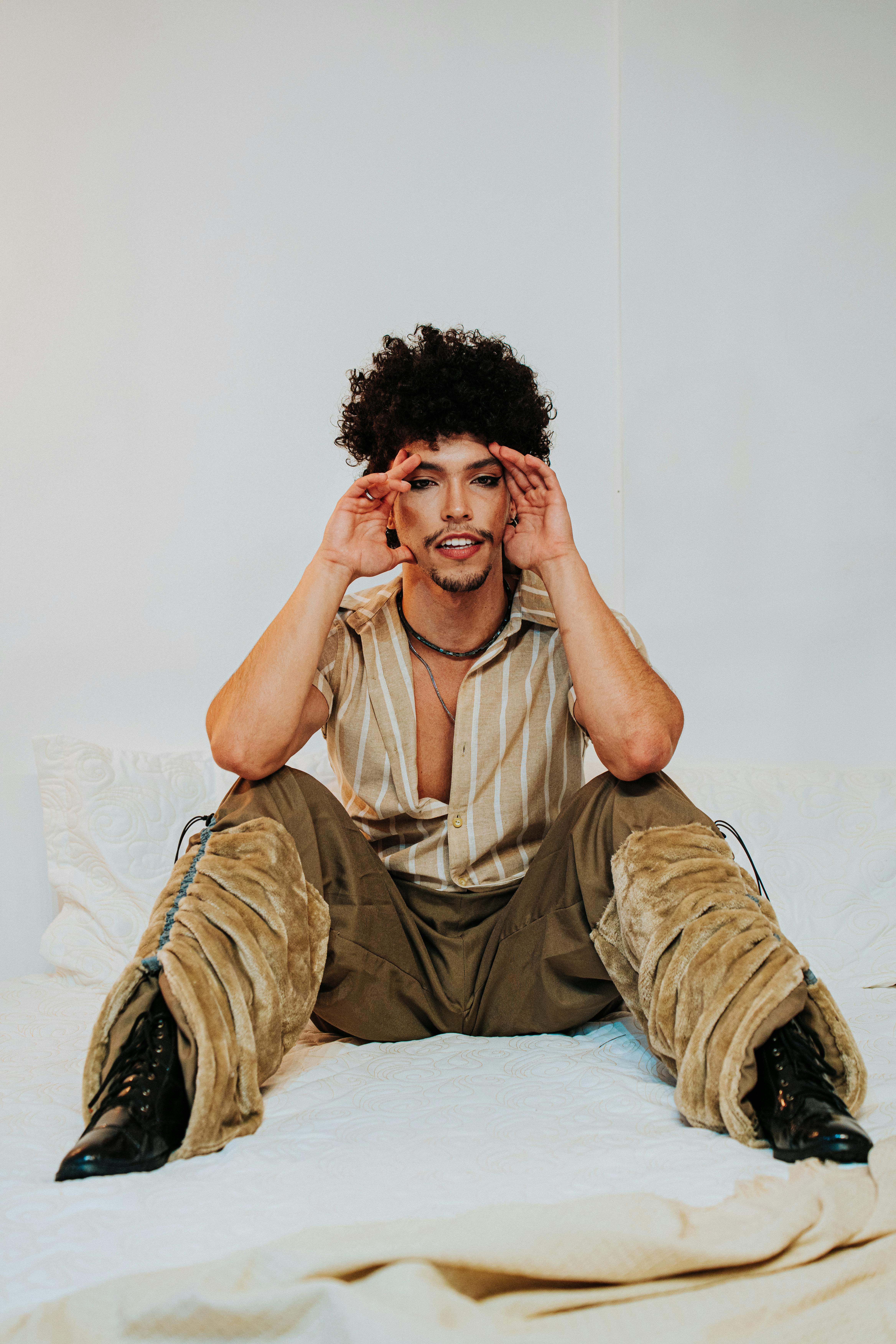Stylish young man with curly hair sitting confidently on a bed in a minimalist room.