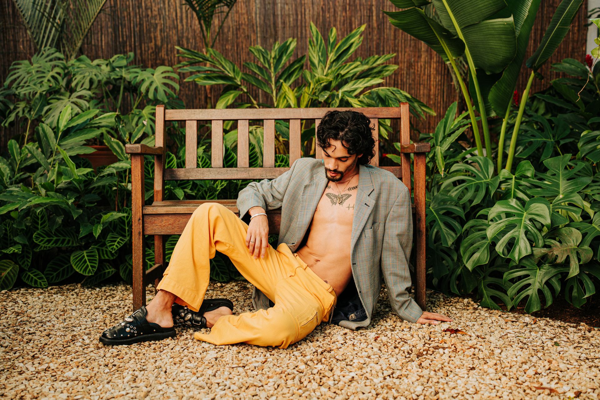 A fashionable man relaxes on a wooden bench surrounded by lush tropical plants in a garden setting.