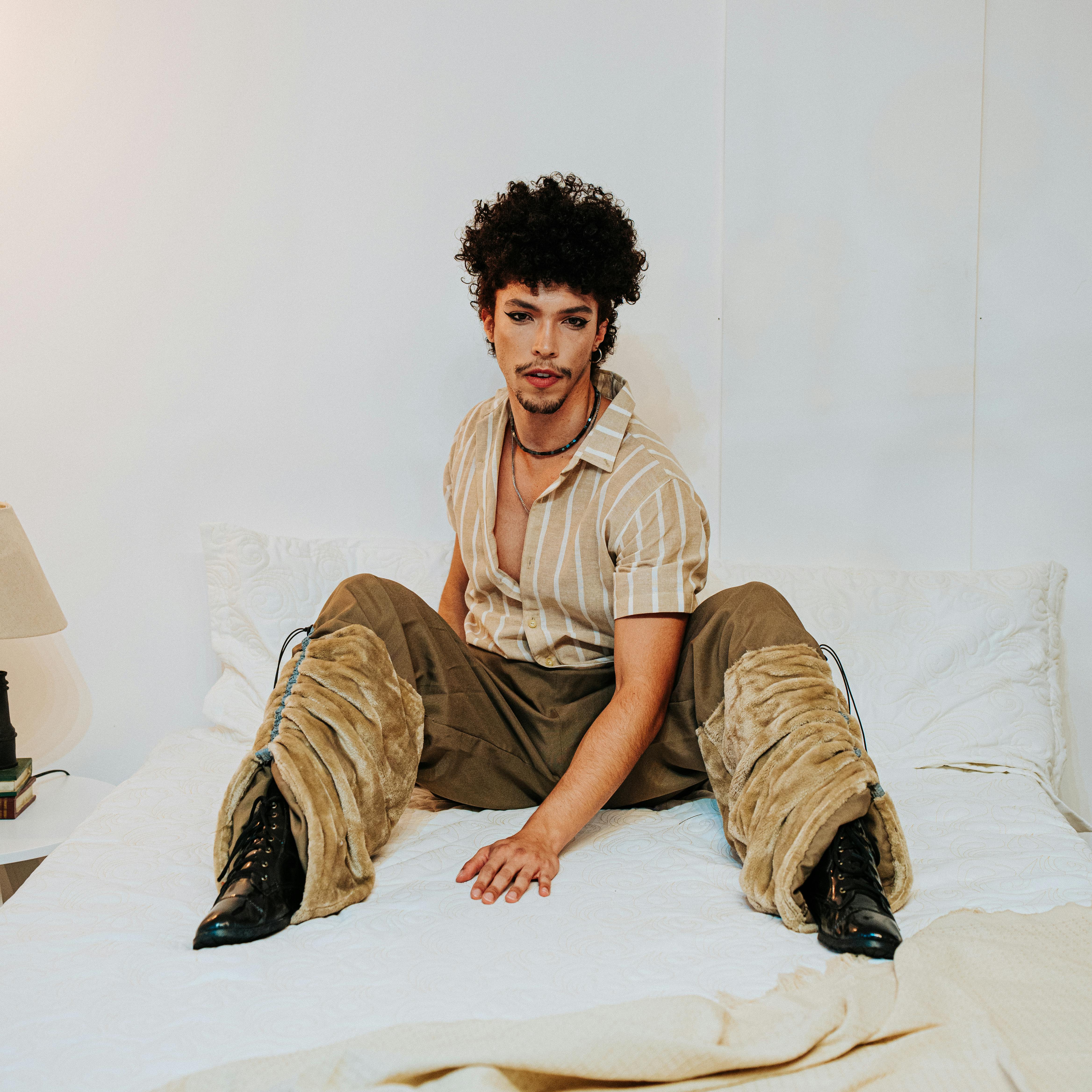 A fashionable man sits on a bed indoors wearing trendy clothes, exuding modern style.