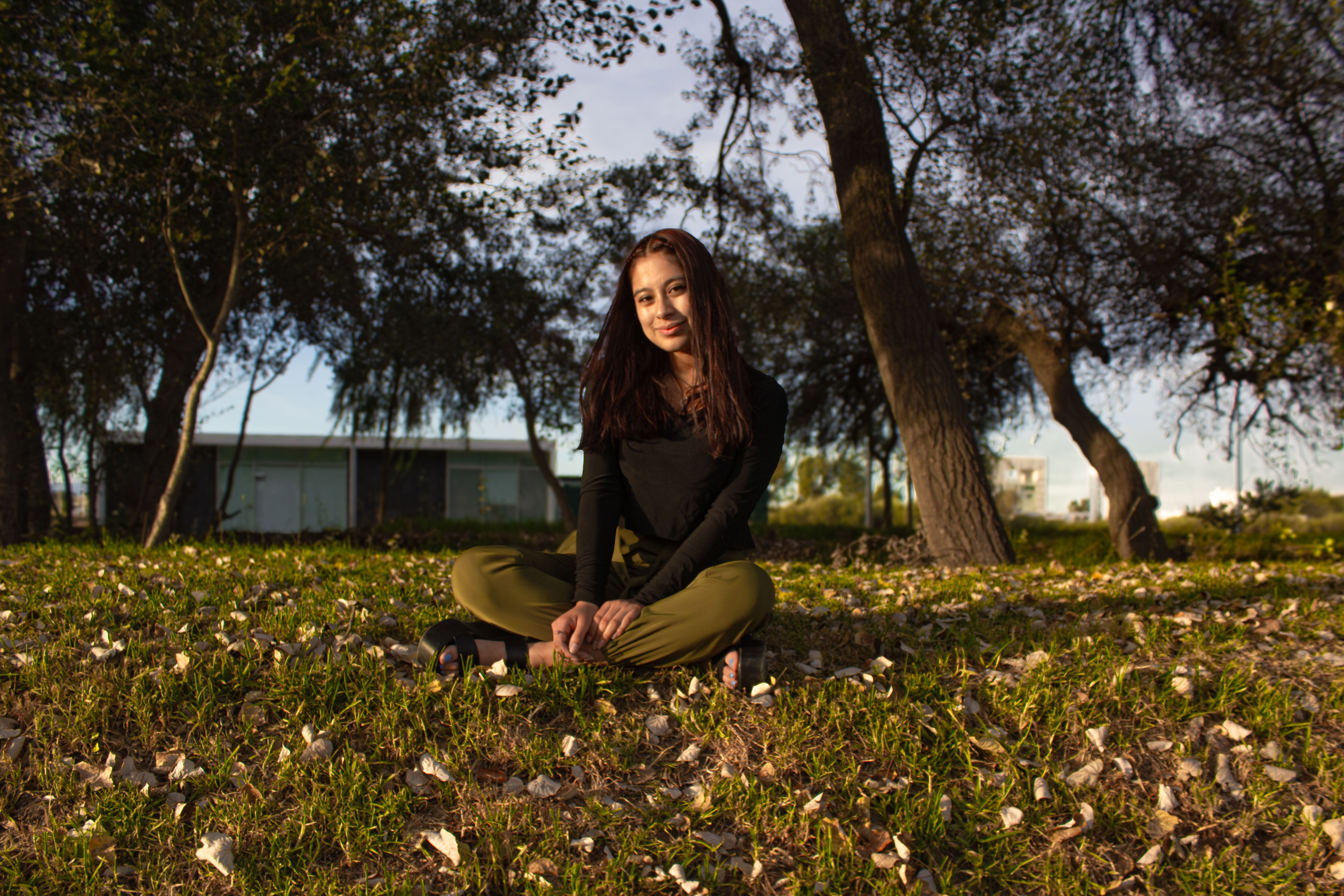 A young woman sits calmly on fallen leaves surrounded by trees in a sunny outdoor setting.