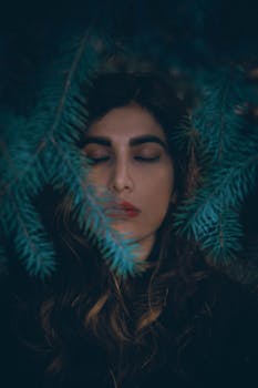这幅艺术特写肖像描绘了一位闭目女子，四周环绕着郁郁葱葱的冷杉绿枝