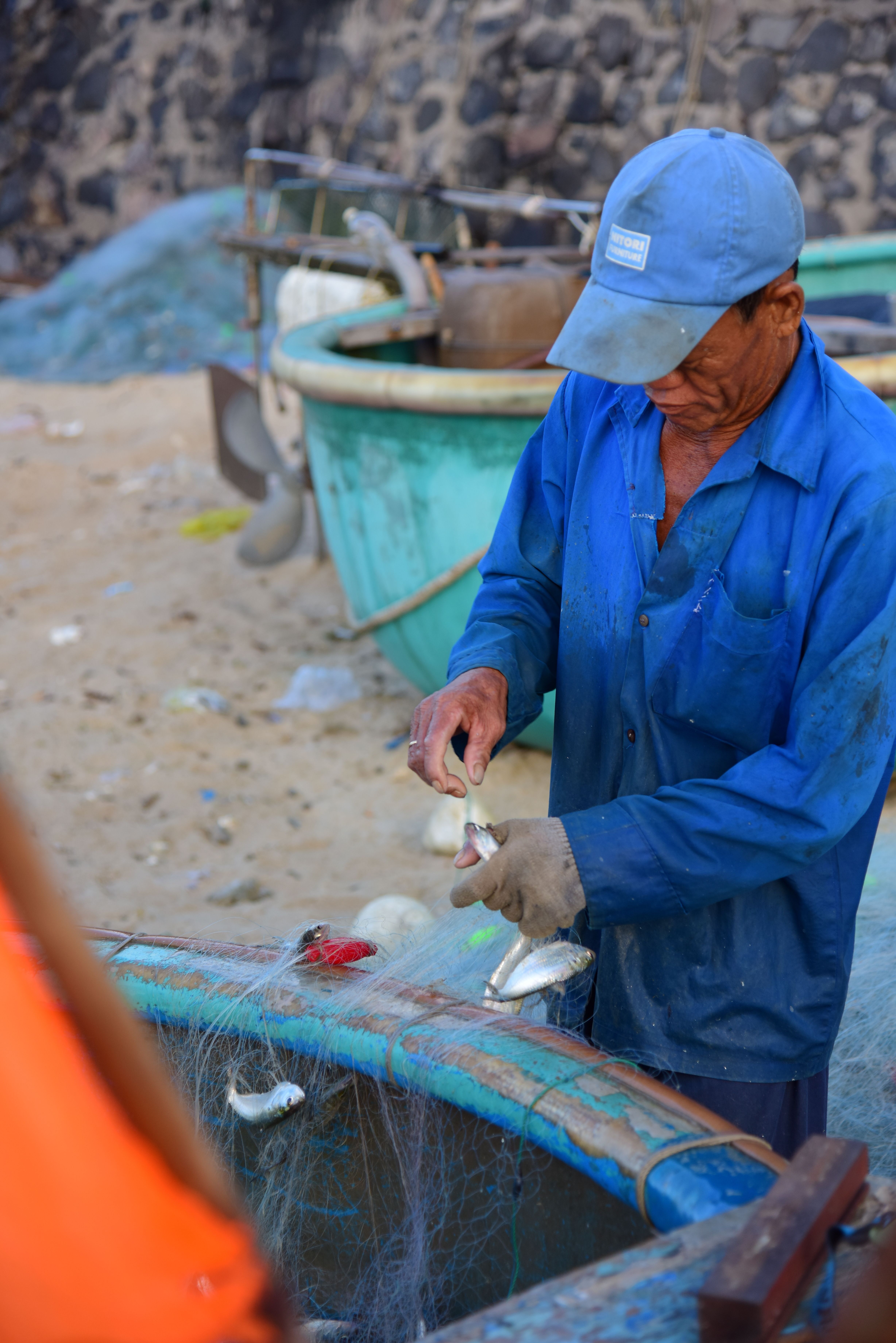A fisherman cleans freshly caught fish with nets and boats in the background. Captured outdoors.