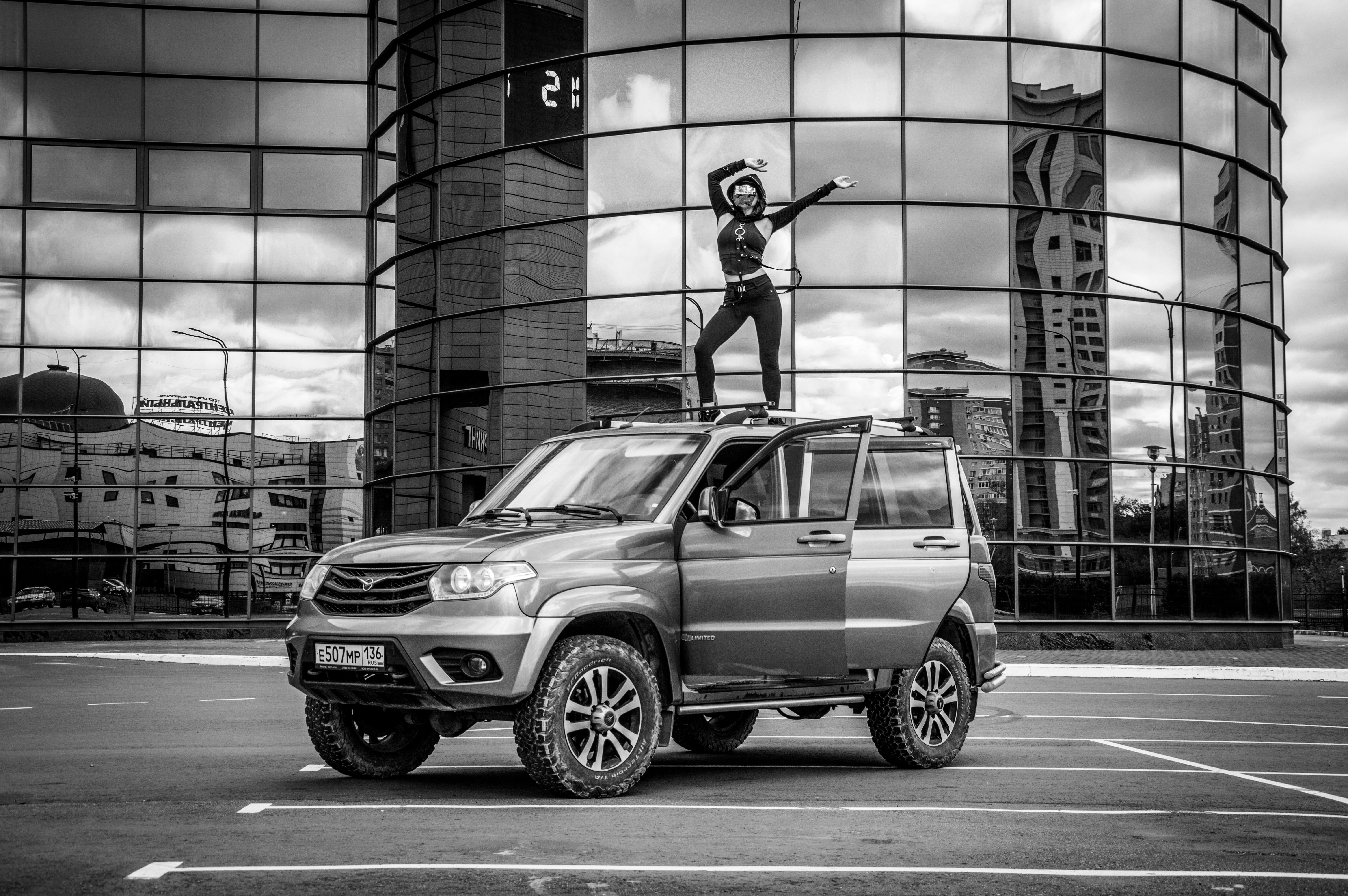 Black and white image of a person striking a dynamic pose on an SUV in a cityscape.