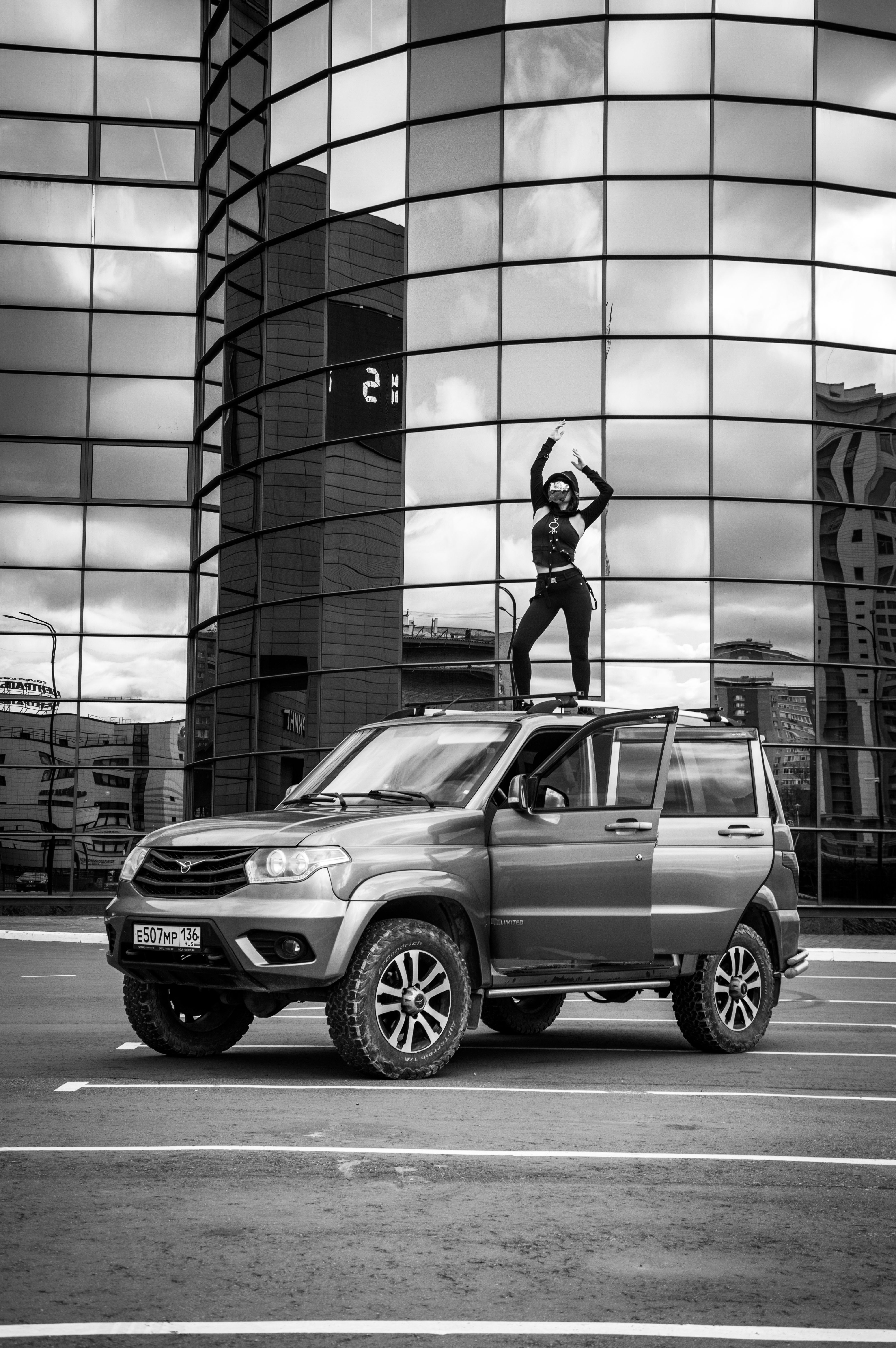 Black and white image of a person dancing on an SUV with a glass building backdrop, exuding urban energy.