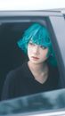 Young woman with striking teal hair gazing thoughtfully from a car window.