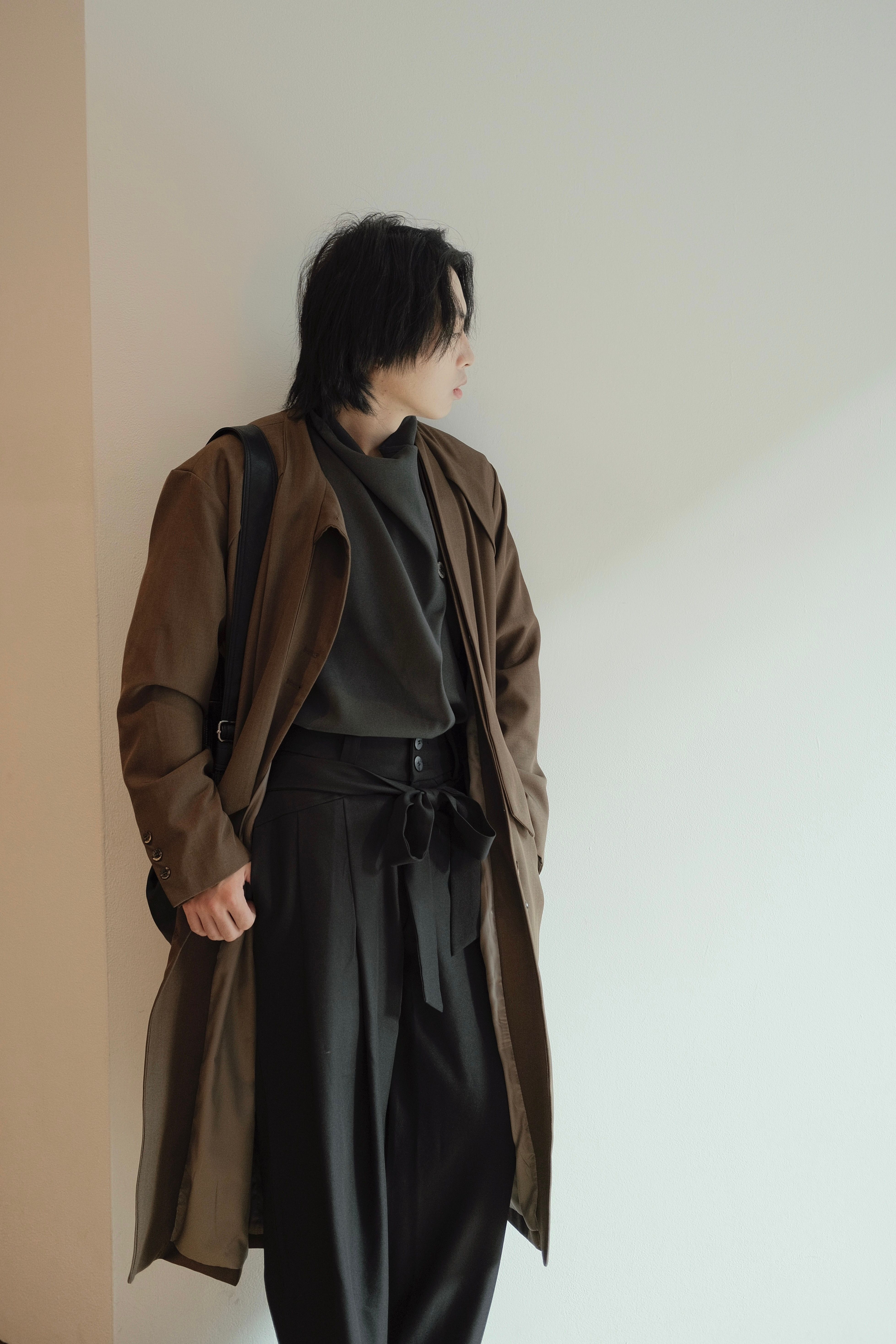 Fashionable man leaning against a white wall with a brown coat and black attire.