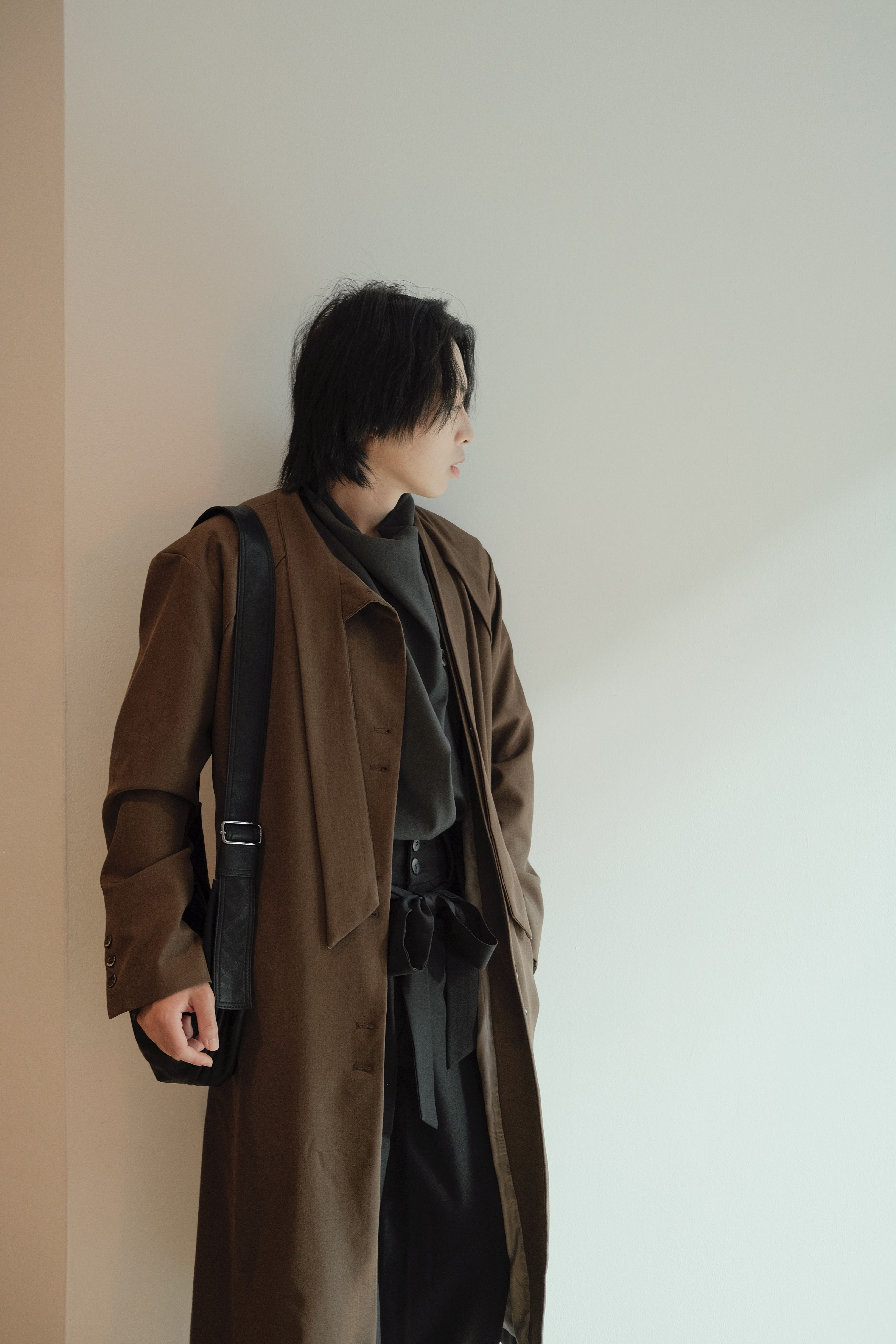 Young Asian man in fashionable brown coat posing against a minimalist indoor background.